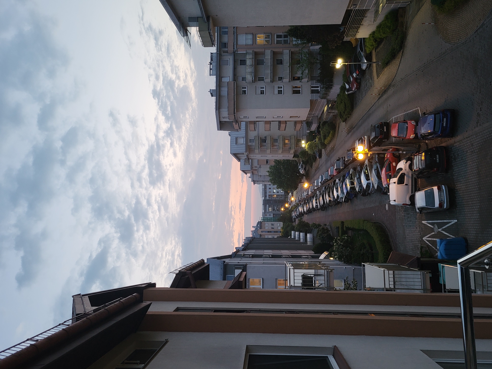 The image depicts an urban scene at dusk. Buildings line both sides of a street filled with parked cars. Streetlights are illuminated, casting a yellow glow on the parked vehicles and the road surface. The sky above is a mix of light blue and white clouds, with hints of orange and pink near the horizon, suggesting the sunset. The perspective is from an elevated position, possibly a balcony, overlooking the street and the buildings. Details of the buildings include windows, balconies, and varied architectural styles.