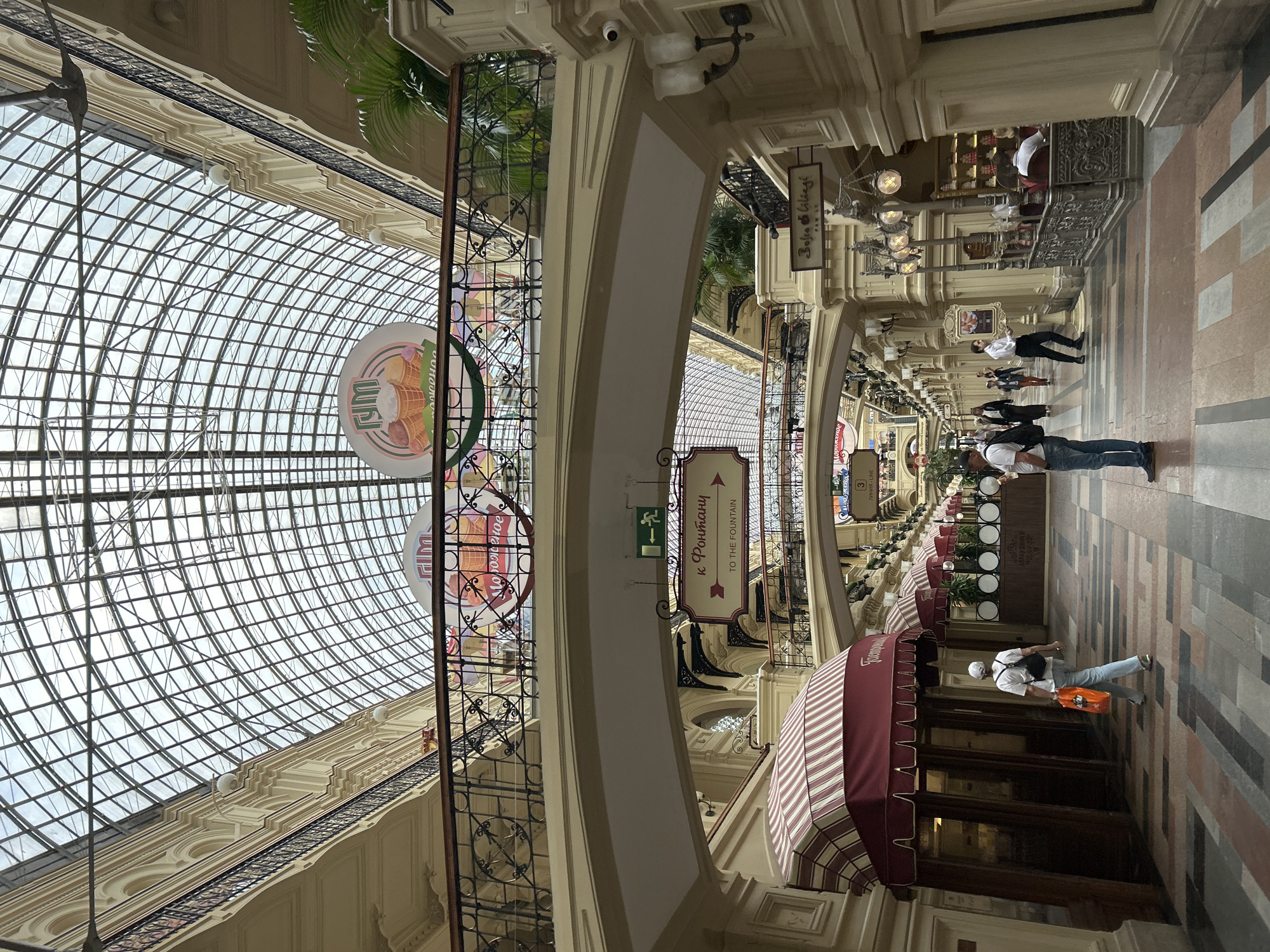 The image shows an interior view of a large, ornate building with a glass ceiling. The scene appears to be a shopping arcade or department store, likely in Moscow, Russia, given the visible Russian text.
Several people are walking through the arcade. In the foreground, a man in a white t-shirt, blue jeans, and a white baseball cap walks away from the camera, carrying an orange bag. Another person, wearing a white shirt and dark pants, walks towards the camera further down the arcade. Other individuals are visible in the background, some appearing to be shoppers and others possibly staff.
The architecture is grand, with arched walkways, decorative moldings, and ornate lamps. The curved glass ceiling allows natural light to illuminate the space. There are various shops or stalls lining the arcade, some with awnings. One awning is striped in red and white.
Visible text includes signs in Russian. One sign with an arrow reads "к Фонтану" which translates to "to the Fountain," with an English translation "TO THE FOUNTAIN" below it. Another sign shows "ЛУКОВЫЕ ЛИНИИ" which translates to "Onion Lines." There is also a circular sign with an image of ice cream cones and Russian text.
The time of day appears to be daytime, as suggested by the natural light. The weather outside is not discernible, but the interior is well-lit. The overall impression is one of a busy, elegant public space.