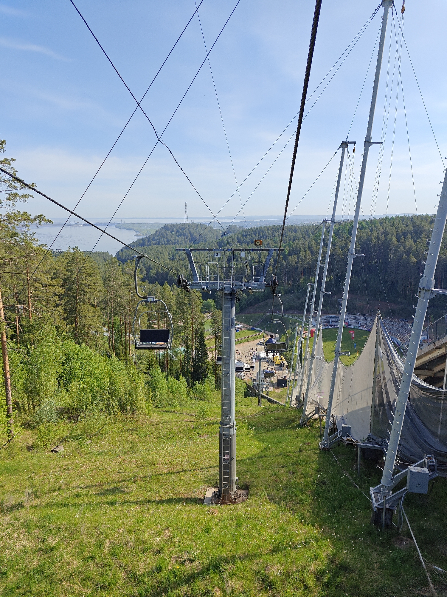 The image depicts a chairlift in Прикамский, Russia. The chairlift ascends a grassy slope, with trees and a body of water in the background. A ski jump with safety netting is visible on the right side of the slope. The sky is blue with some clouds.
