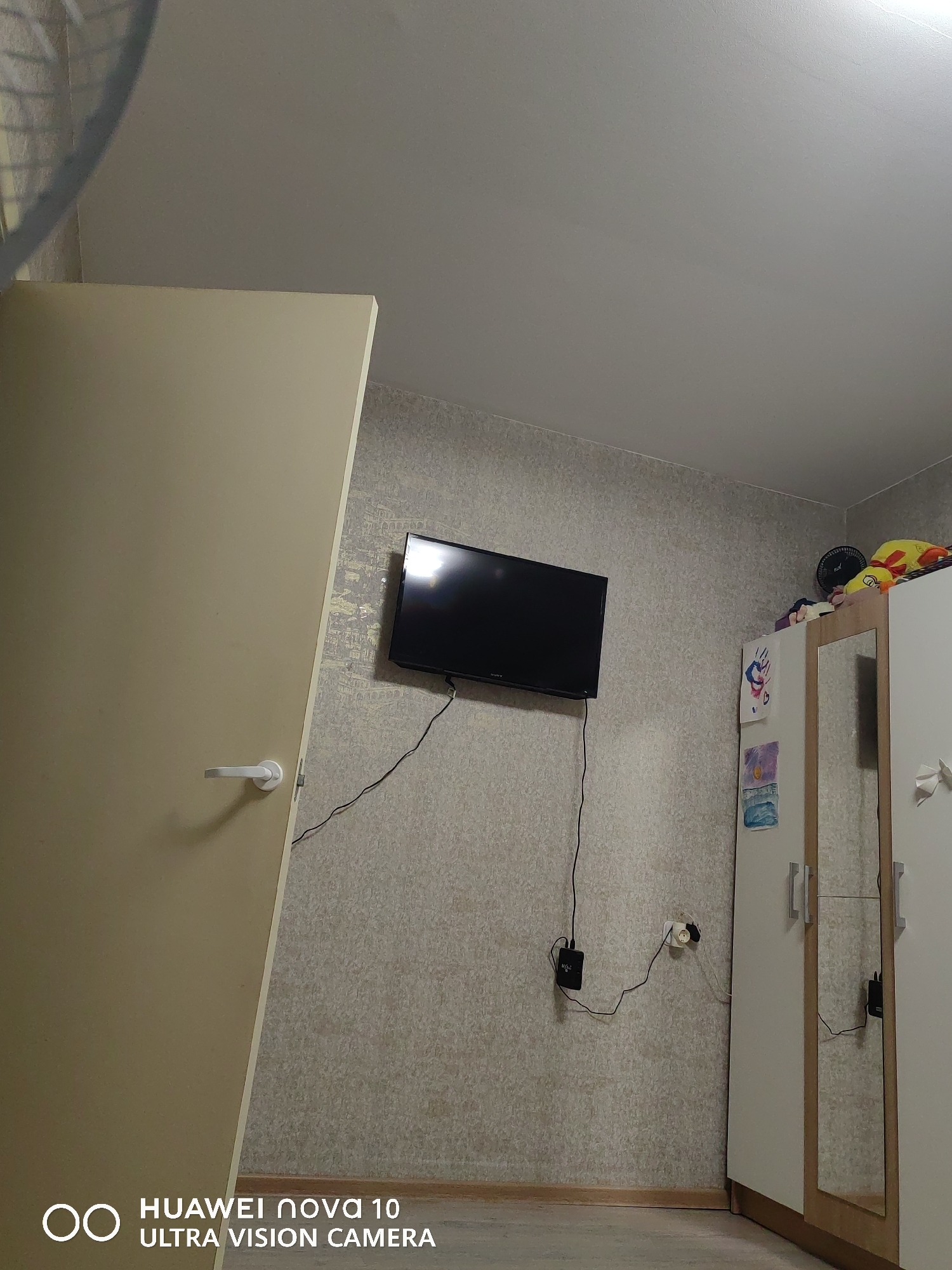 An indoor scene, likely a bedroom or living space, is captured from a low angle, looking towards a wall with a television and a wardrobe. On the far left, a light beige or off-white door, possibly slightly ajar, with a white handle, occupies a significant portion of the frame. The wall behind the door and TV is covered in a light-colored, textured wallpaper with a subtle pattern.
A black flat-screen television is mounted on this wall. Cables run from the TV downwards to a black power adapter or device, which is connected to a white electrical outlet near the floor. To the right, a white wardrobe with light wood trim features a mirrored door and a plain door. Several colorful plush toys and a small black fan are stored on top of the wardrobe. On the left side of the wardrobe, several children's drawings or artworks are taped.
The floor is light-colored, appearing to be laminate or wood. The ceiling is plain and light gray. A blurred, curved object, possibly part of a ceiling fan or a standing fan, is partially visible in the top left corner. No people are present in the image. The lighting is bright, suggesting daytime or well-lit indoor conditions. A "HUAWEI nova 10 ULTRA VISION CAMERA" watermark is visible in the bottom left corner.