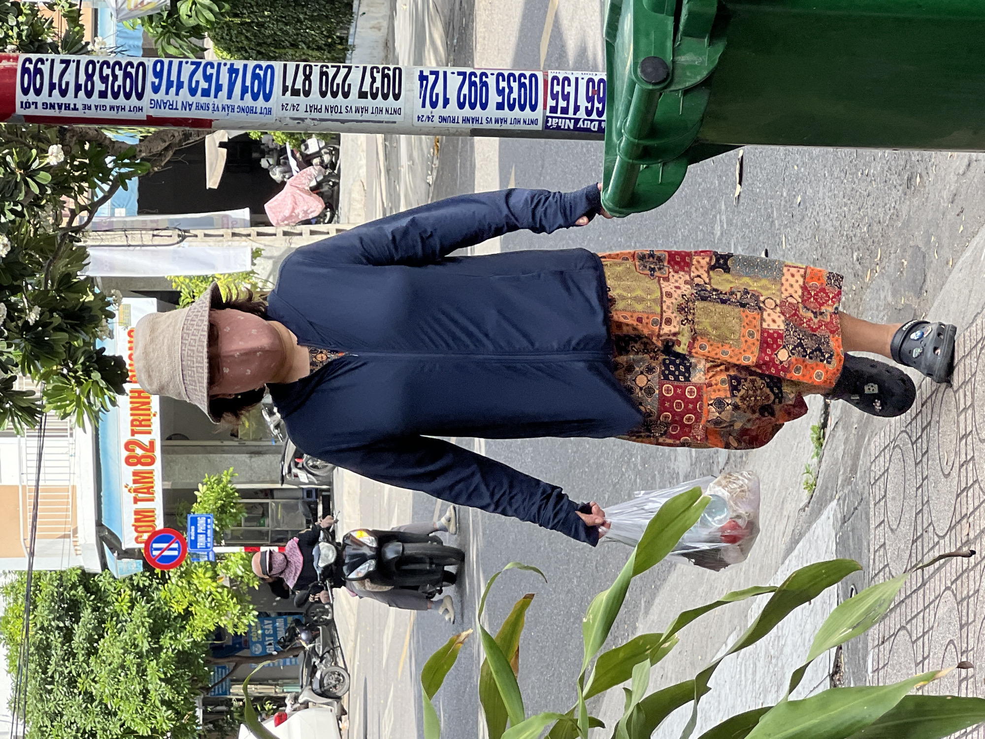 A woman walks on a sidewalk in Nha Trang, Vietnam. She is wearing a light-colored bucket hat, a blue zippered jacket over a patterned dress, and black Crocs. A pink patterned face mask covers her nose and mouth. She carries a clear plastic bag in her right hand. To her left, a green trash bin is visible. In the background, traffic signs, a building with a blue awning reading "CƠM TẤM 82 TRỊNH PHONG", and parked motorcycles are present. A man is riding a motorcycle in the street.