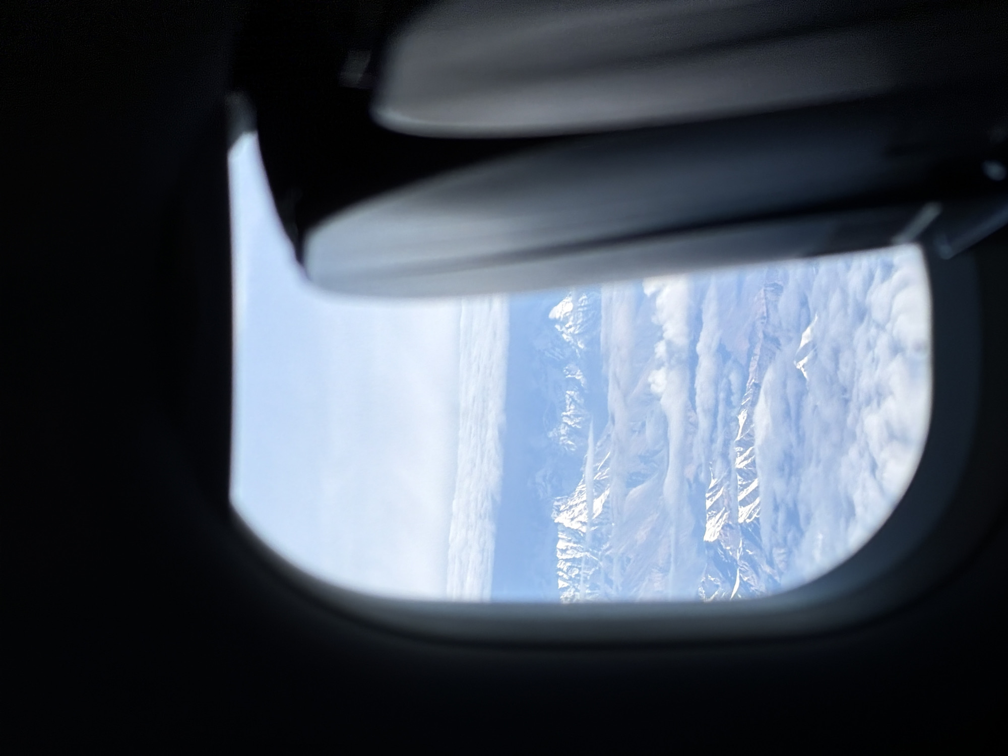 An aerial view from an airplane window shows snow-capped mountains peeking through clouds. In the foreground, the dark interior of the airplane, likely the seatback and window frame, partially obscures the view. The mountains appear rugged and vast, with some peaks showing sharp, angular ridges. The sky is a bright, clear blue, with some wispy clouds scattered around the mountain range. The lighting suggests it is daytime, likely mid-morning or early afternoon, given the brightness of the sky. The image captures a scenic and remote landscape from a high altitude.