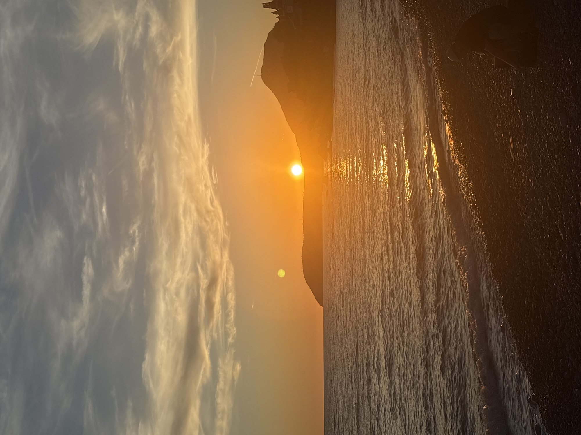 The image depicts a serene sunset scene at a beach in Bar, Montenegro. The sun, a brilliant orange orb, is setting behind a silhouetted hill, casting a warm golden glow across the sky and the water. Wispy clouds are scattered across the upper half of the frame, tinged with the colors of the sunset. The sea is calm, with gentle waves lapping at the pebble shore. In the foreground, on the right side of the image, a lone figure is seated on the beach, facing away from the camera and towards the sea, appearing to be contemplating the sunset. The beach is covered in dark pebbles. The overall atmosphere is peaceful and tranquil.