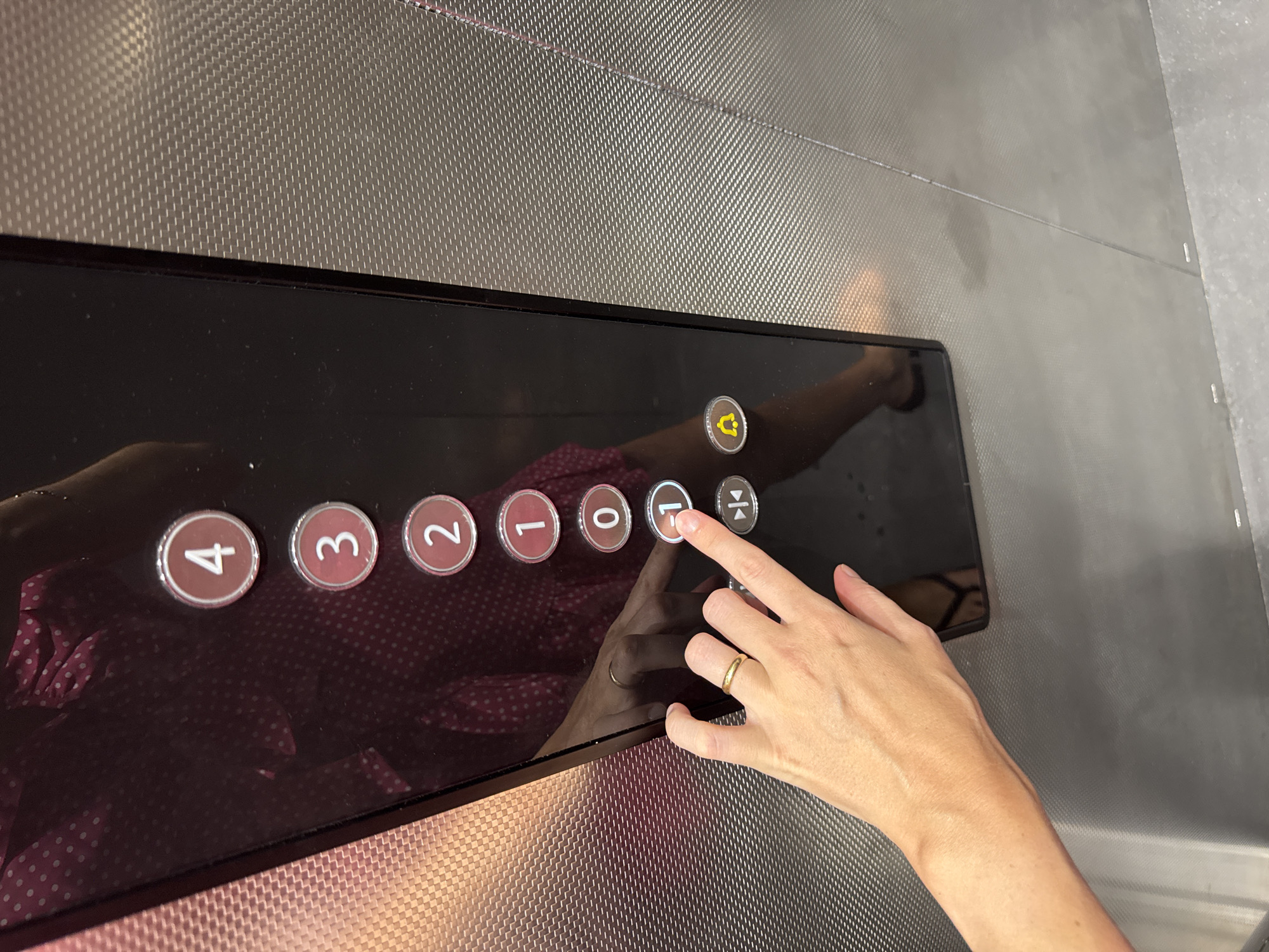 In Isola delle Femmine, Italy, a person's hand with a gold ring is pressing the button labeled "-1" on an elevator control panel. The elevator panel is black and features illuminated circular buttons for floors 4, 3, 2, 1, 0, and -1. Below the floor buttons are two additional icons: one resembling an open and closed door, and another of a bell. The surrounding elevator wall has a textured metallic finish. A reflection on the dark surface of the panel shows a person wearing a dark pink polka dot dress.