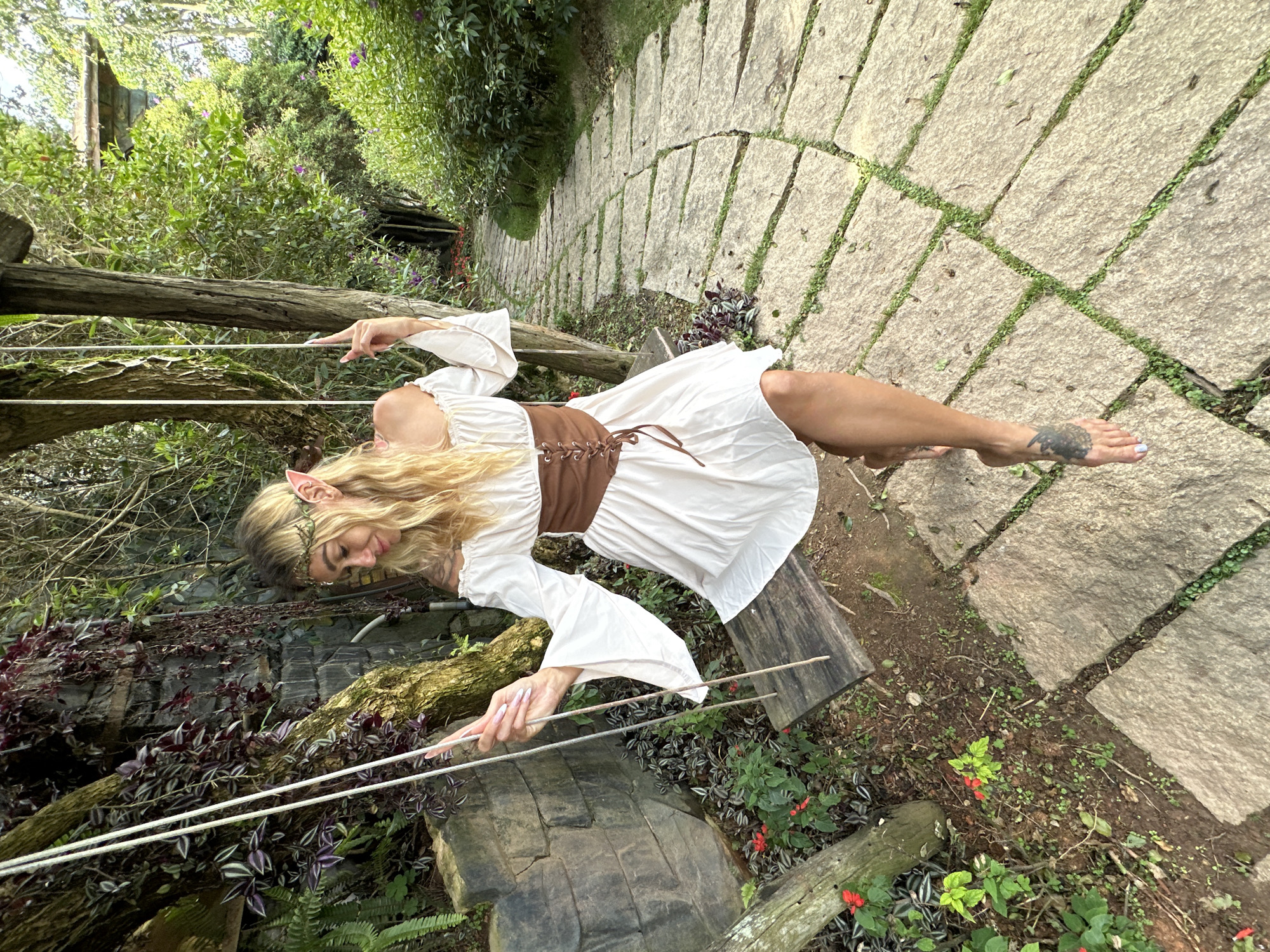 The image shows a woman dressed in a white, loose-fitting peasant blouse with flared sleeves and a brown corset. She has long blonde hair and is wearing an elf-like ear accessory. The woman is outdoors, in what appears to be a garden or wooded area with a stone pathway. She is reaching out with both hands, seemingly touching or interacting with two taut white cords that are suspended horizontally. Her body is angled, with her left leg extended, revealing a tattoo on her left foot. The setting includes lush green foliage, trees, and a stone walkway with grass growing between the stones. In the background, there is a glimpse of what might be a rustic structure, possibly wooden. The overall impression is one of a fantasy or whimsical theme. The lighting suggests it is daytime, and the abundant greenery indicates a mild climate. The location is identified as Da Lat, Vietnam. There is no visible text in the image.