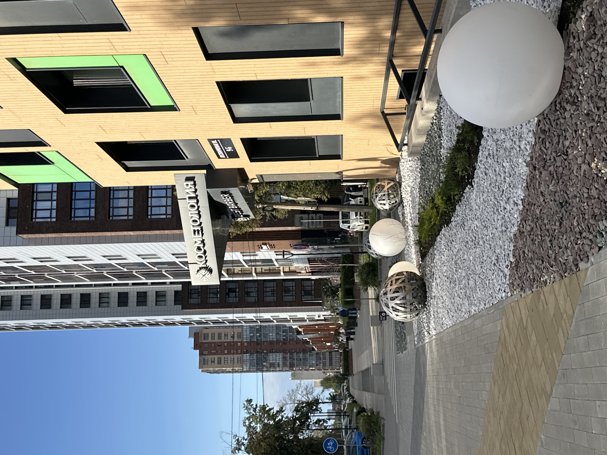 A modern cityscape is captured on a sunny day with a clear blue sky. The foreground features a paved walkway bordered by decorative gravel beds. Large, spherical white lights and a metallic orb sculpture are placed within these beds. To the right, a contemporary building with a beige, textured facade and black-framed windows stands prominently. Green panels add a splash of color to some window surrounds. A metal railing runs alongside steps leading down from the building. In the mid-ground, a street runs parallel to the walkway, with parked cars and a few pedestrians walking along the sidewalk. Further in the background, a row of multi-story apartment buildings rises, showcasing a variety of architectural styles and colors. The overall scene suggests a well-maintained urban environment with a mix of residential and commercial structures.