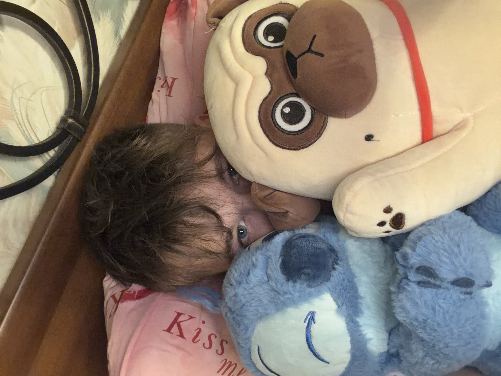 In a bedroom, a person, likely male, peeks out from behind two plush toys: a blue Stitch character on the left and a light brown pug on the right. The person's eyes are visible, and their hair is dark and messy. The plush toys are large and occupy most of the frame. The background includes a wooden headboard and a pink pillow with red lettering that appears to say "Kiss me." The overall impression is of a cozy, personal space. This image was not taken in Saint Petersburg, Russia.