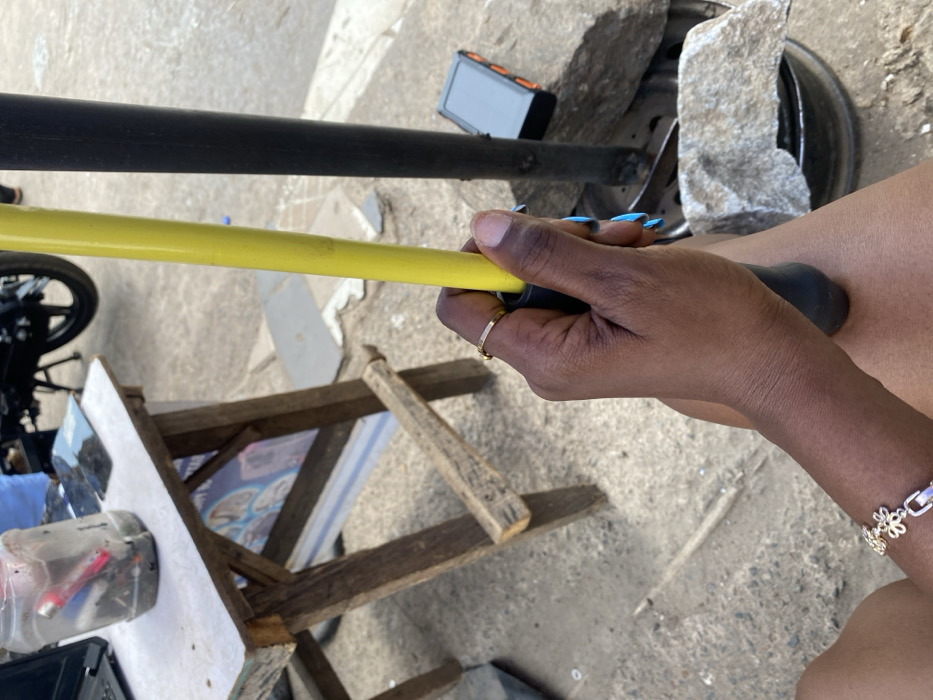 The image depicts an outdoor street scene in Ibadan, Nigeria, characterized by a mix of commercial activity and utilitarian elements. In the immediate foreground, a dark-skinned human hand grasps a yellow pole with a black rubber grip. A thin gold-colored ring is present on the ring finger, and fingernails are painted light blue. A silver-colored bracelet with interconnected flower-shaped links adorns the wrist. The person's bare upper thigh is visible below the hand, suggesting a seated or resting posture.
To the left of the frame, a weathered wooden table with a white laminated top serves as a makeshift workspace. On the table, a clear plastic jar, likely a repurposed bottle, contains miscellaneous items including a red flashlight and other small objects, possibly tools or debris. A black laptop is partially visible on the far left of the table, and several pieces of cracked or disassembled glass, resembling phone screens, are laid flat. Behind the table, a partially visible banner or poster displays graphics that appear to be electronic components.
Further into the scene, a black vertical pole stands to the right of the yellow pole, possibly another support structure. Near its base, a compact black and orange electronic device, possibly a power bank or portable modem, rests on the ground. A gray rock sits atop what appears to be a discarded black tire rim, also on the ground. The ground surface consists of rough, light-colored dirt or concrete. In the background, a motorcycle tire and a portion of a black motorcycle are visible on the right, and a person's legs wearing light blue pants are partially visible behind the table. The overall lighting is bright, indicative of daylight.