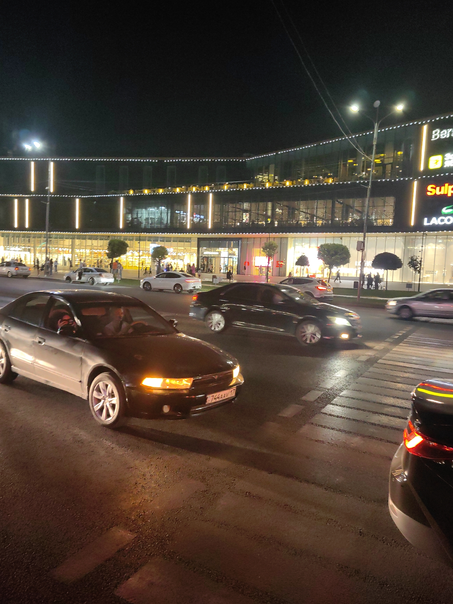 At night, a dark-colored sedan with its headlights illuminated is driving on a wet asphalt road, crossing a pedestrian crosswalk. The car's license plate is visible and reads "744AAV 17". Behind this car, a white sedan is seen turning left. In the background, a multi-story building with extensive glass windows is lit up with a facade decorated with vertical white and yellow lights. Signs on the building read "Bershka" and "Sulpak", suggesting it is a shopping center. Several trees are planted in front of the building. The sky is dark, indicating nighttime. The overall scene captures a street view in a city environment.