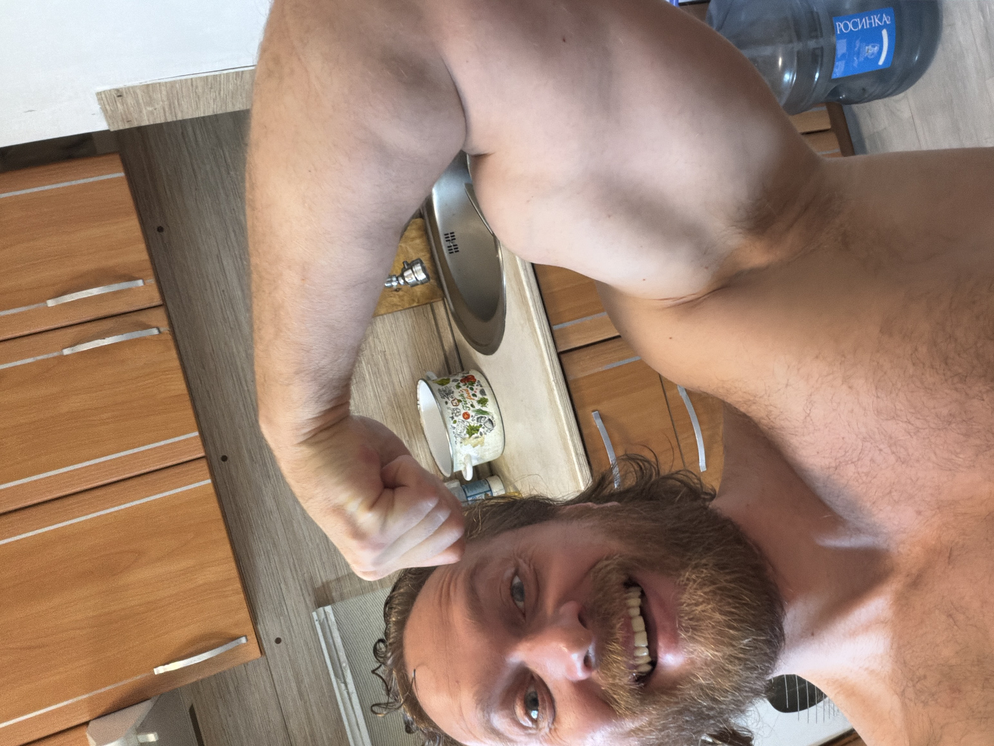 The image is a vertical, close-up selfie of a shirtless man, appearing to be taken in an indoor setting like a kitchen or utility room. The man, likely in his 30s or 40s, has a full reddish-brown beard and medium-length dark, curly hair. He is smiling broadly, revealing his teeth, and looks directly at the camera with a cheerful and energetic expression.
He is flexing his left bicep, which is prominently displayed towards the viewer, indicating a show of strength or a playful pose. His upper body is muscular, and he has fair skin with some body hair on his arms and chest.
The background reveals parts of a domestic space. To the left, wooden cabinets or drawers with silver horizontal handles are visible. A metal kitchen sink with a faucet is partially obscured behind the man's head and shoulder. On the counter near the sink, a white ceramic mug with colorful floral or fruit patterns can be seen. Further back, to the right, a large, clear plastic water bottle with a blue label is present. The label clearly shows the Cyrillic text "РОСИНКА" (ROSINKA), which is a common brand of bottled water in Russia. The floor appears to be a light-colored wood or laminate. The overall lighting suggests an interior shot, possibly during the day or under artificial illumination. The visible text on the water bottle serves as a strong location cue, consistent with Saint Petersburg, Russia.