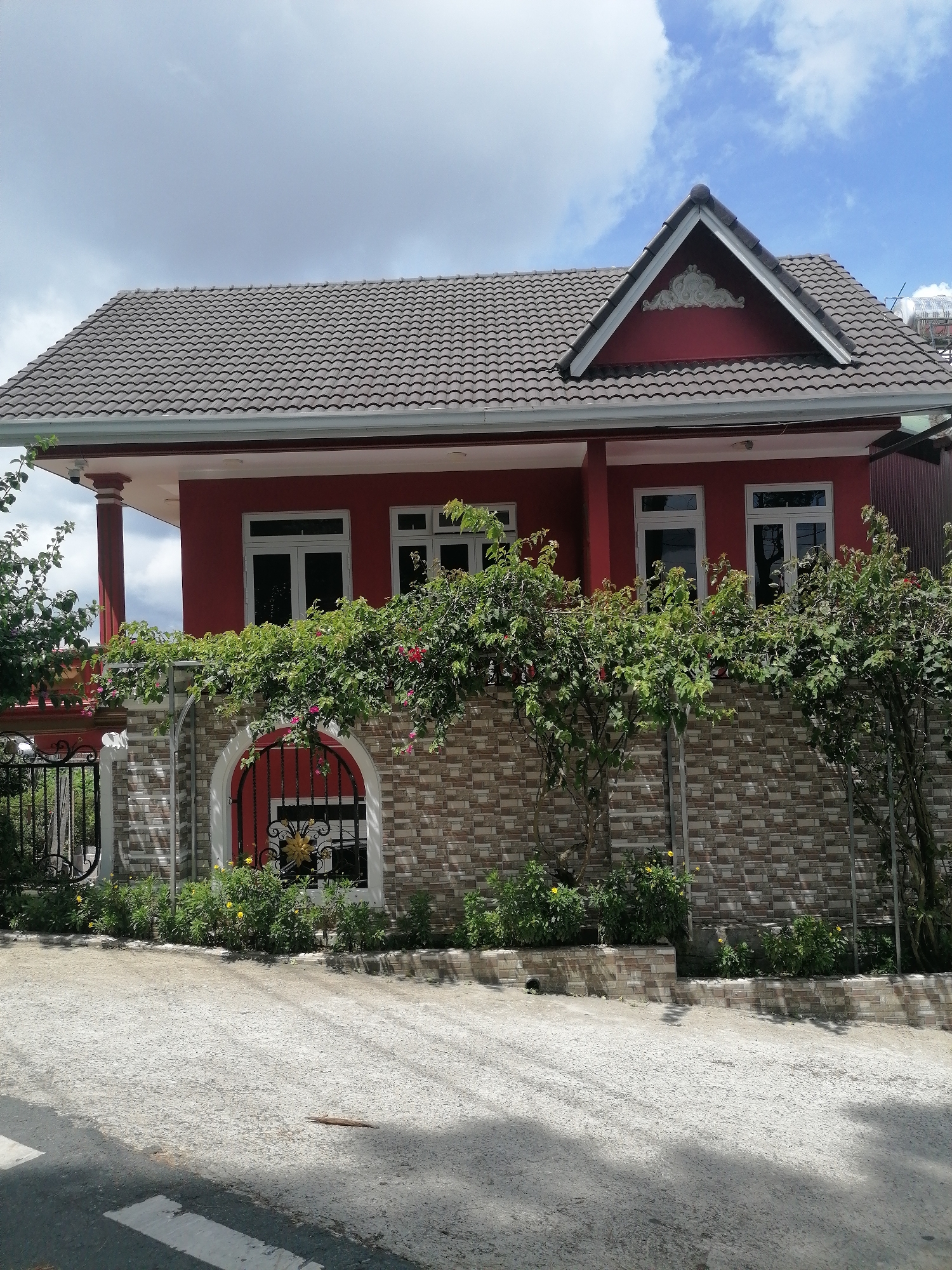The image shows a red two-story house with a gray tiled roof in the city of Da Lat, Vietnam. The house has white-framed windows and a prominent gable with decorative molding. A stone wall with a decorative arched entrance separates the house from the street. Lush green foliage, including flowering vines, grows over the wall and around the property. A black metal gate is visible on the left side of the wall. The ground in front of the house is paved with concrete and appears to be a sloped driveway or street. The sky is partly cloudy with blue patches. No people are visible in the image.