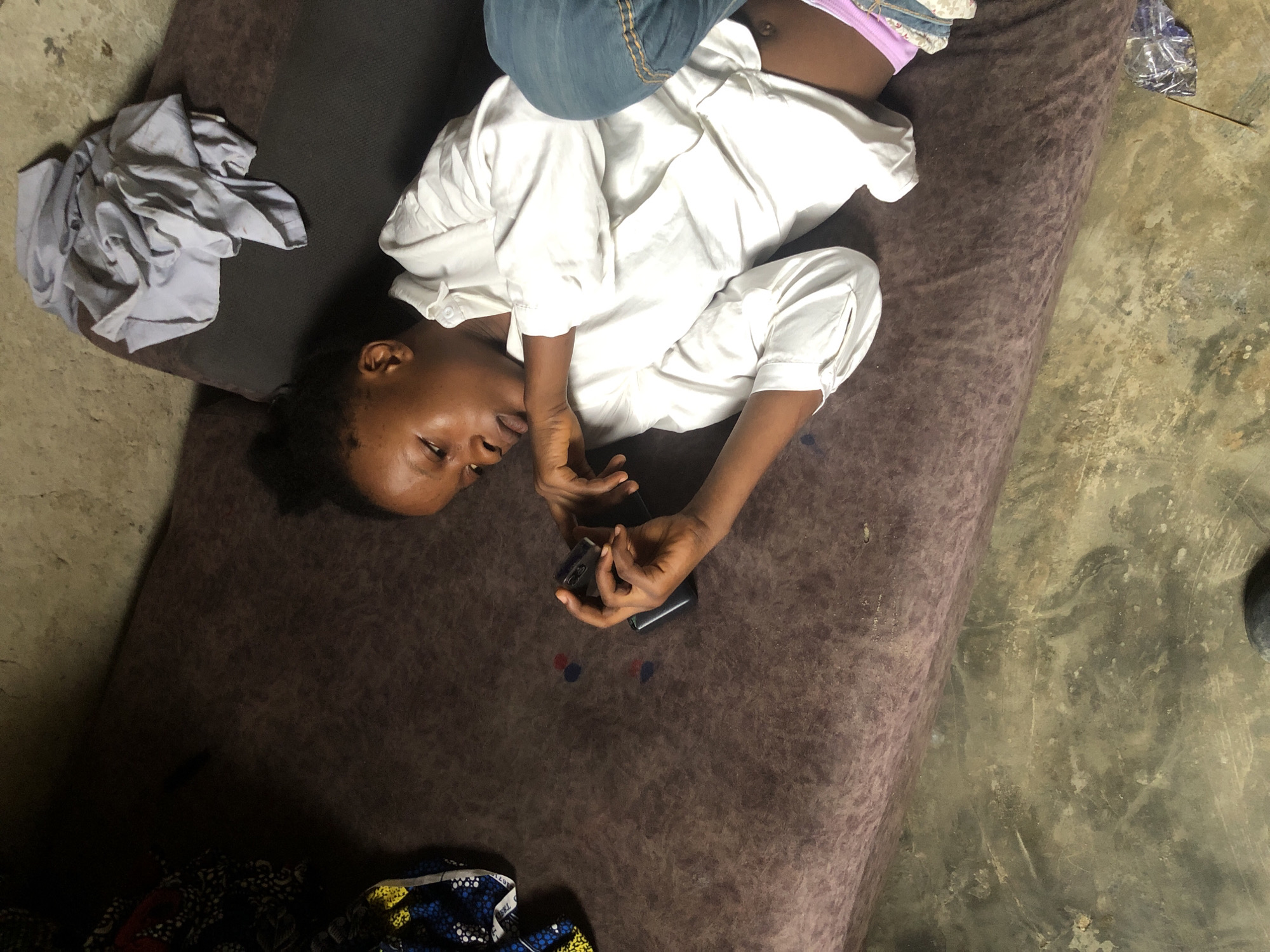A young Black female lies on a dark brown mattress. She is wearing a white button-down shirt, blue jeans, and pink underwear. She is looking at a black cell phone in her hands. Her hair is dark and pulled back. There are two small circular red and blue ink stains on the mattress to her left. To her left and slightly above her head are pieces of colorful fabric. To her right and above her is a pile of light gray fabric. The mattress appears to be on a floor made of concrete or dirt. The overall environment appears to be a simple, possibly rural or informal living space. The lighting is dim and appears to come from above. There is no indication of the specific location within Ado, Nigeria.