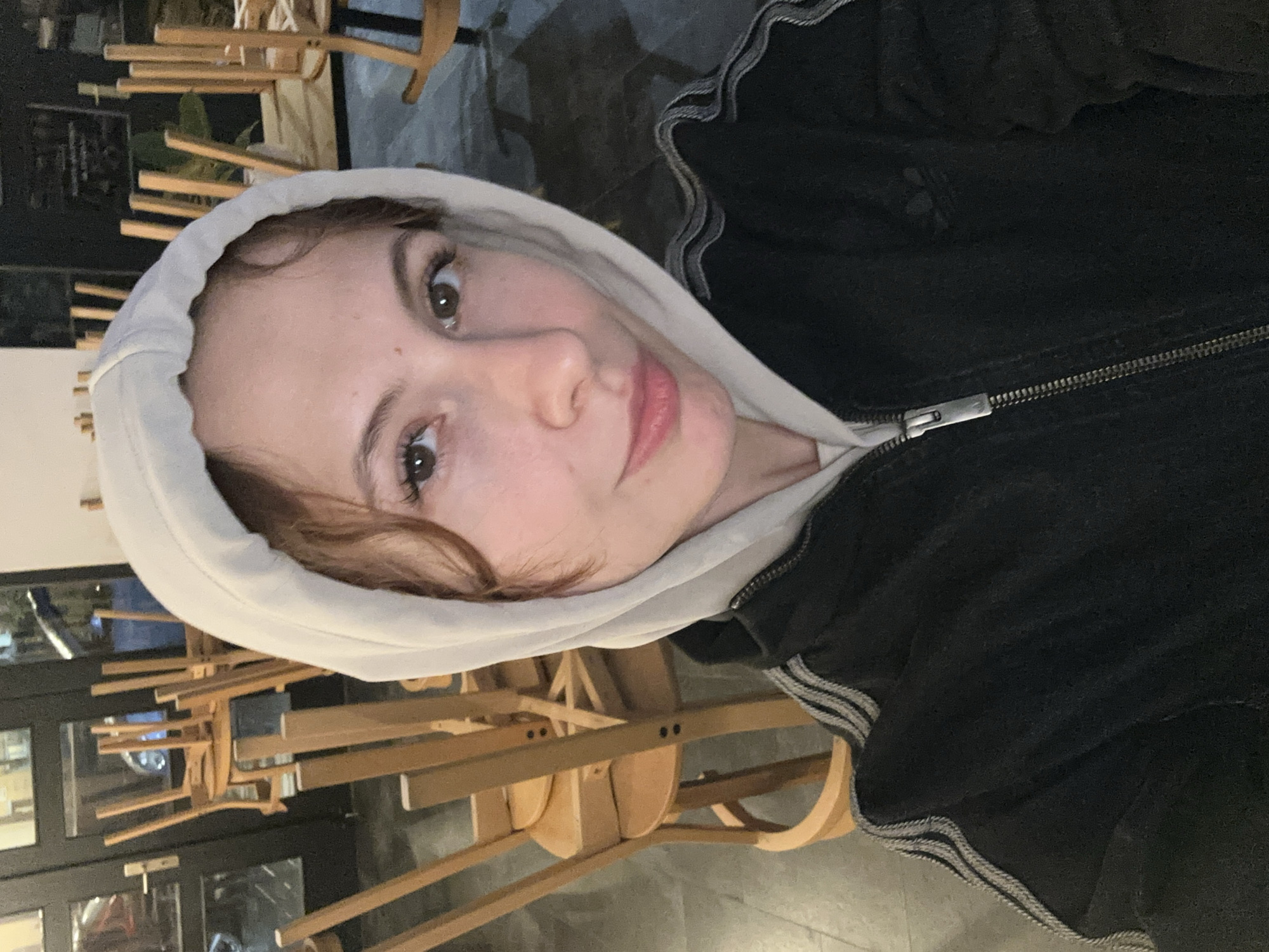 A selfie of a young woman in a white hooded sweatshirt and a black jacket with light gray stripes on the sleeves. She is looking at the camera with a slight smile. The background appears to be a restaurant or cafe, with stacked wooden chairs. Based on the style of the chairs and the general ambiance, it is potentially a restaurant or cafe located in Novi Sad, Serbia.