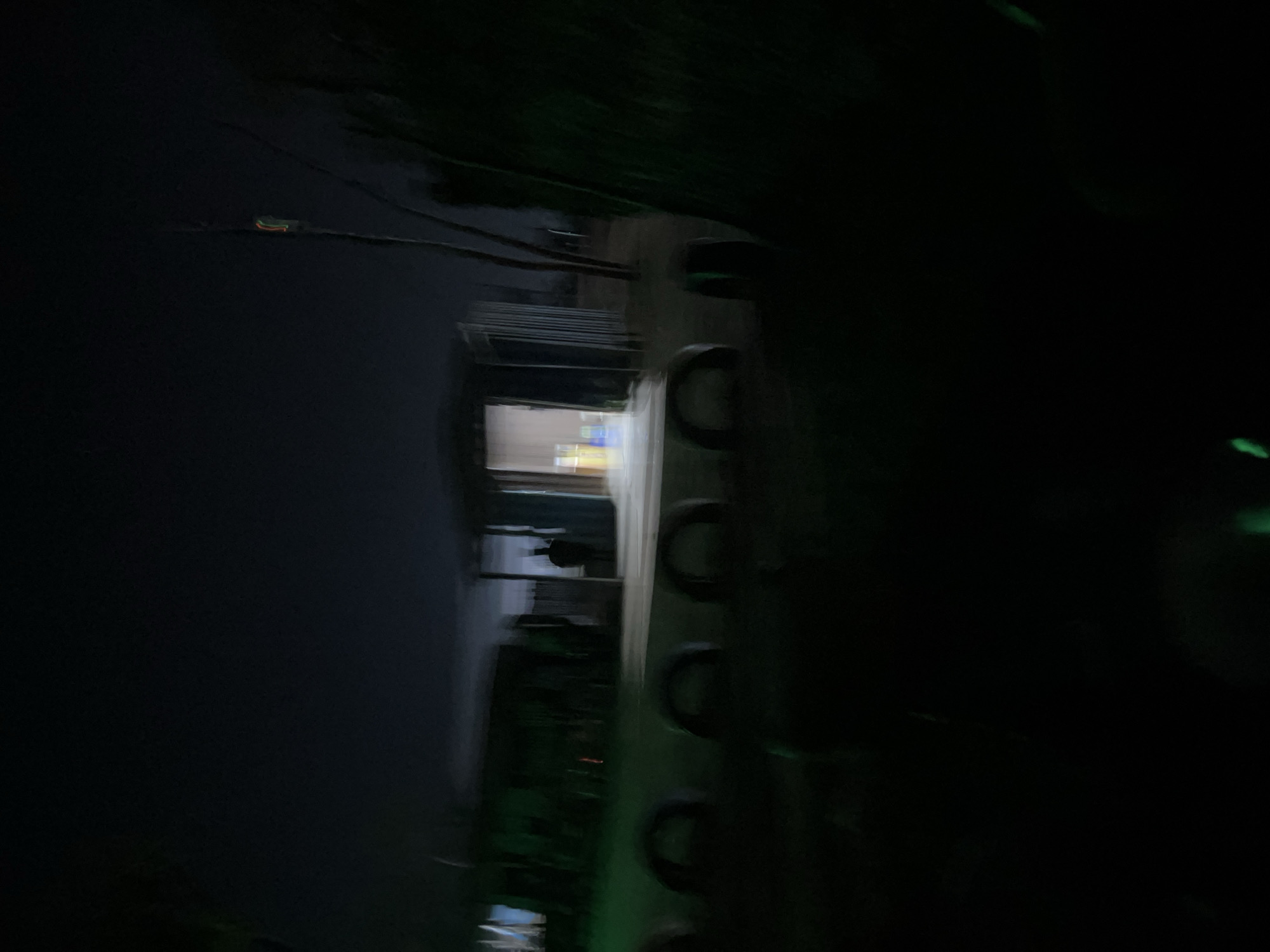 The image depicts a dimly lit outdoor scene at night, likely in a rural or semi-urban area. In the background, a small, open-fronted structure, possibly a shed or makeshift stall, is visible. The interior is illuminated, revealing shelves with what appear to be bottles or containers. A silhouetted figure stands just inside the entrance. In the foreground, a row of what seem to be repurposed tires, cut in half and placed upright, forms a barrier or decorative element. The overall impression is one of a slightly disheveled, possibly abandoned or under-construction site, with sparse lighting adding to a somewhat eerie atmosphere. The location is identified as Howayi, Nigeria.