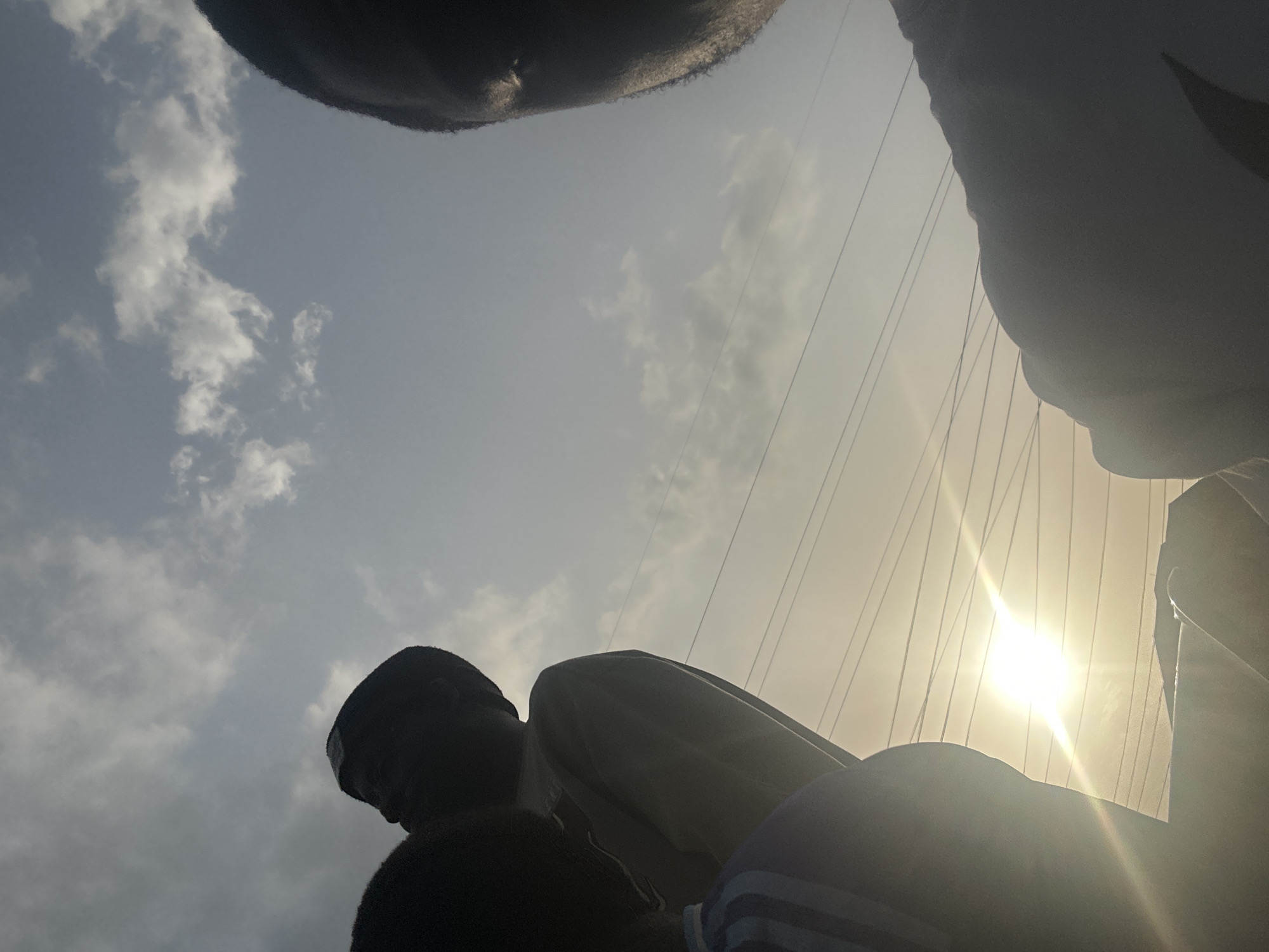 The low-angle shot captures the faces of a group of people standing in Umuode, Nigeria. The sun is shining brightly, positioned near power lines. Clouds are visible in the sky.