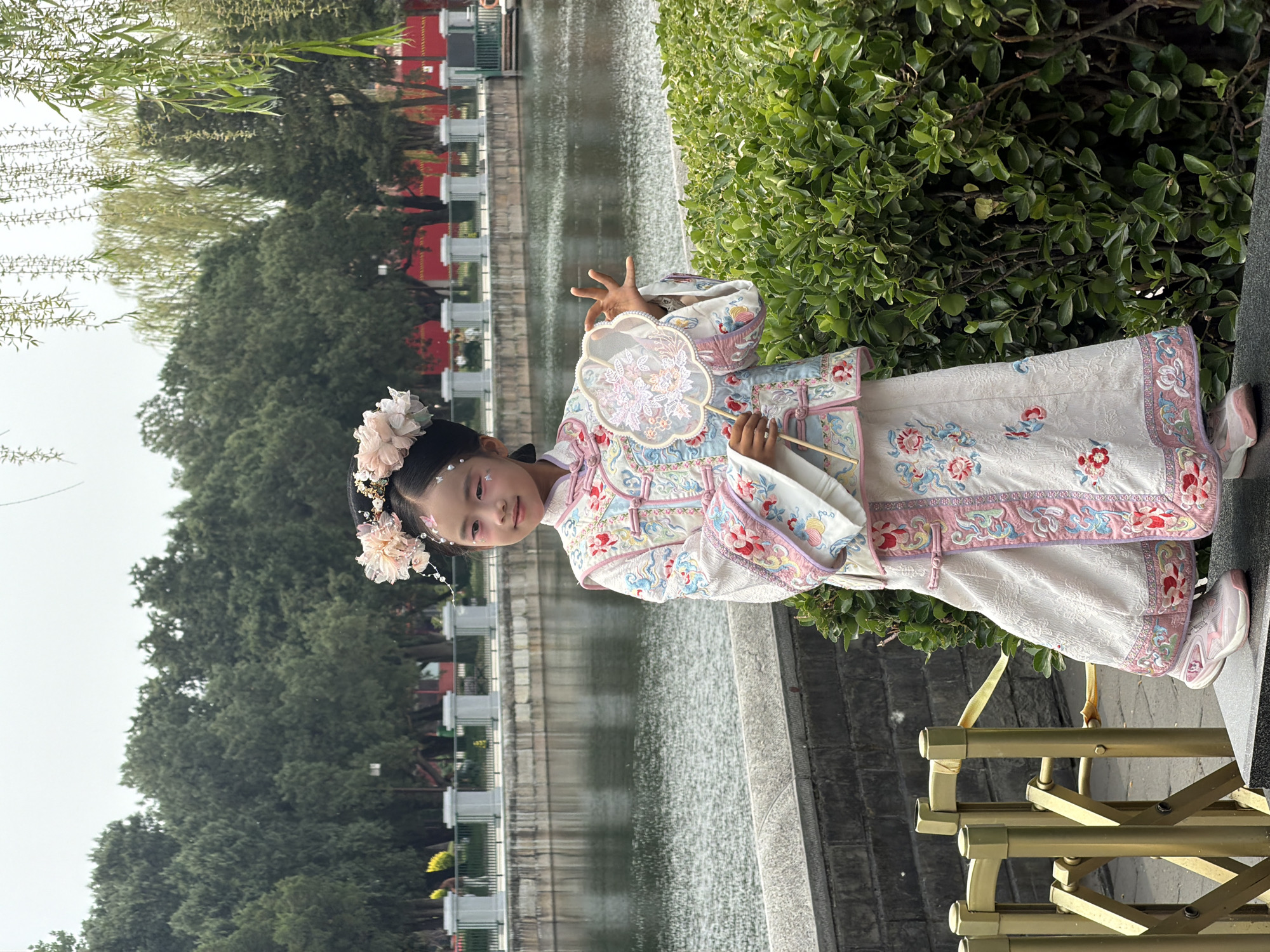 A young girl stands near a body of water, possibly a canal or lake, wearing an ornate, traditional Chinese costume featuring floral patterns and decorative trim. She also has a floral hairpiece and is holding a fan. The environment suggests a park or garden setting with lush greenery and a walkway or barrier in the foreground. The image could be taken in the Dongcheng District, China, given the style of architecture and clothing.