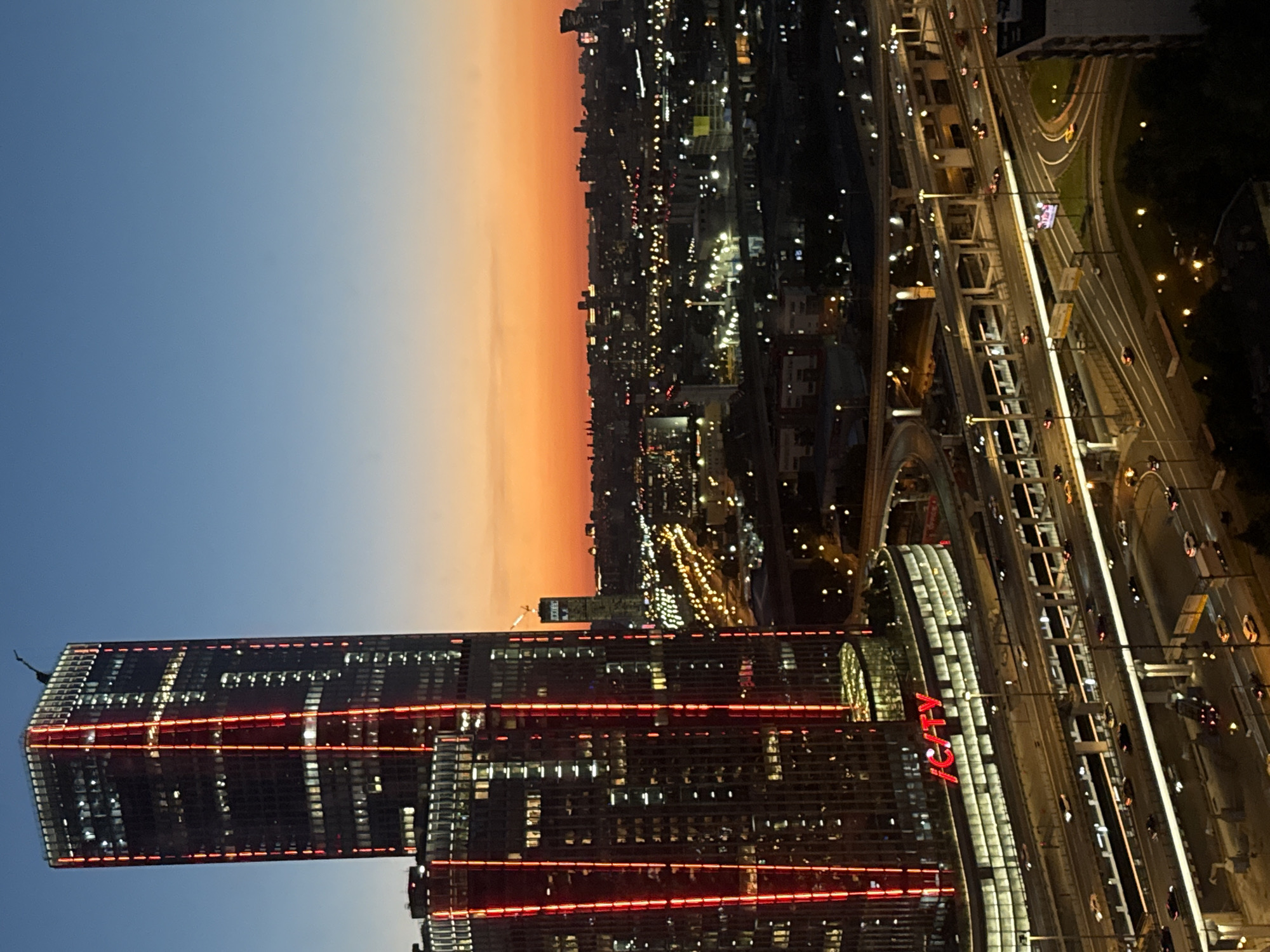 This image depicts a nighttime cityscape with a prominent skyscraper illuminated by red vertical lights. The skyscraper, identified as located in Khimki, Russia, features multiple floors with visible interior lights. Below the skyscraper, a complex network of elevated highways is visible, with cars driving on them and streetlights illuminating the scene. The sky transitions from a deep blue at the top to an orange and yellow gradient near the horizon, indicating dusk or dawn. The surrounding urban landscape consists of numerous other buildings and illuminated lights, suggesting a densely populated area. No people are clearly discernible in the image.