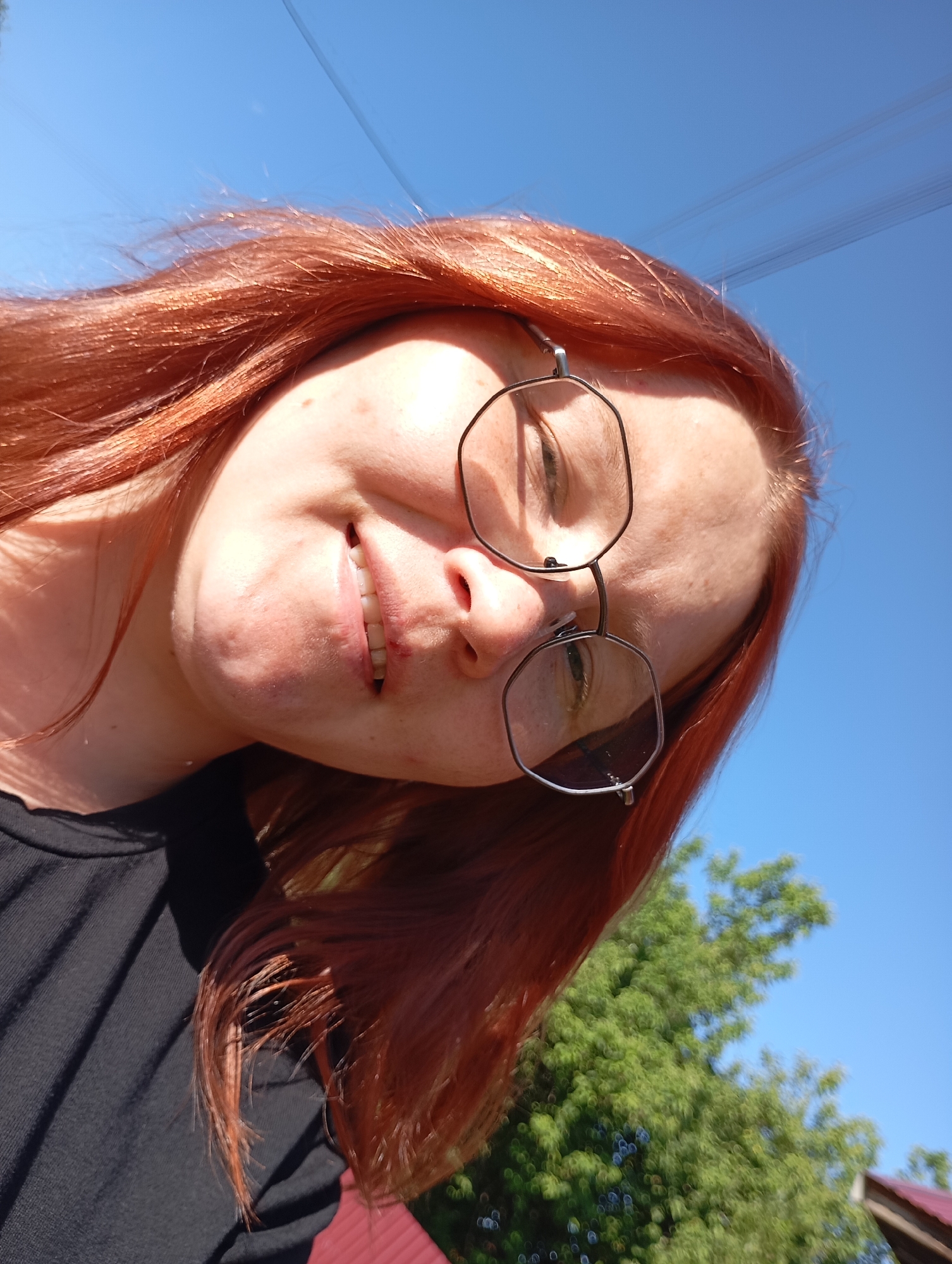 A close-up, low-angle outdoor selfie shows a person with reddish-orange hair and freckles looking upwards. They are wearing black clothing and distinctive octagonal-shaped glasses with thin metal frames. The person's mouth is slightly open, revealing teeth. The background consists of a bright blue sky and green foliage, suggesting a daytime scene in a natural or suburban setting. There are no specific landmarks visible that would definitively place the location in the city of Фоки, Russia. The overall impression is casual and relaxed.