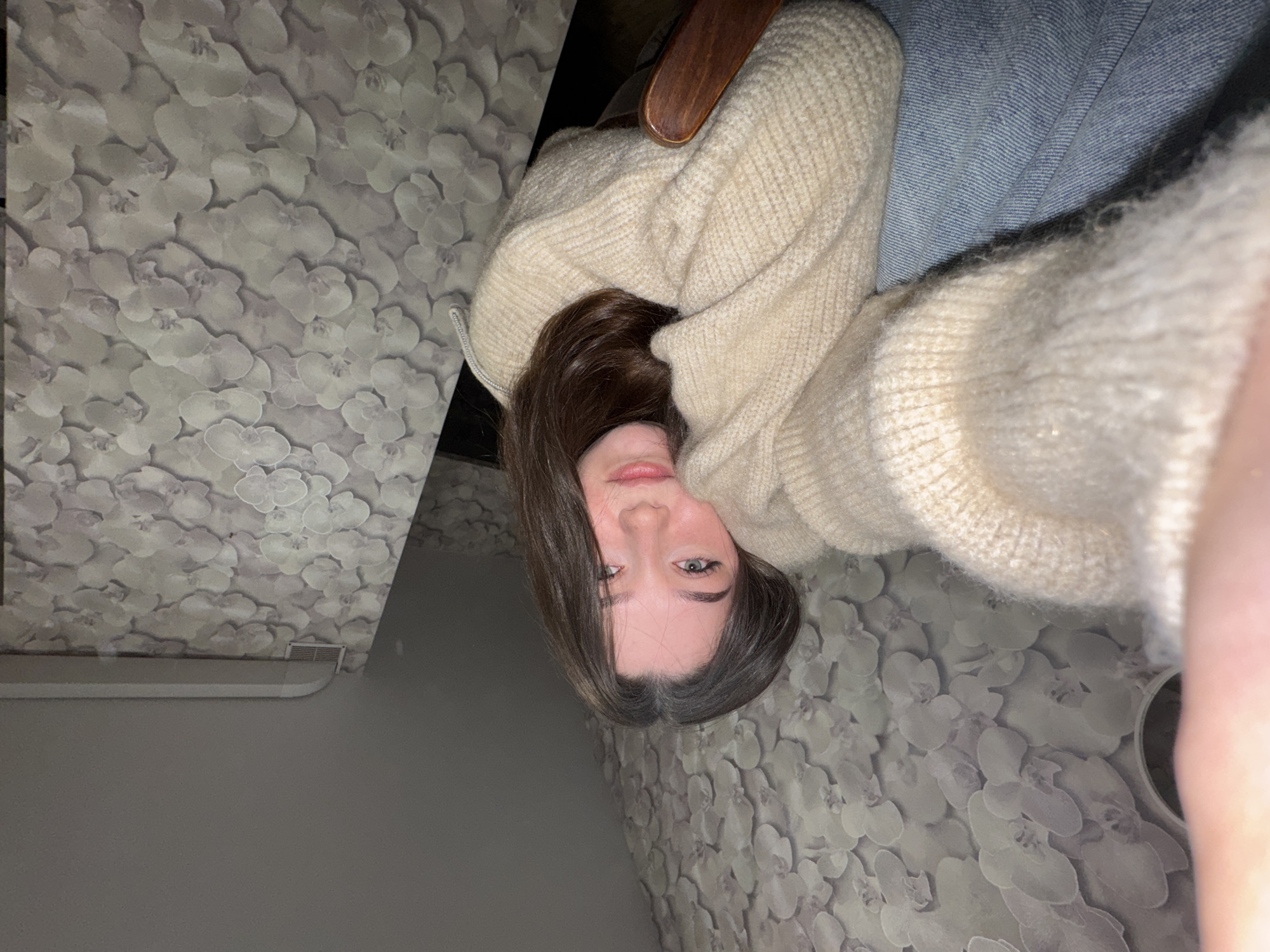 A young Caucasian woman with long, dark brown hair is taking a selfie indoors. She is wearing a beige, knitted sweater and blue jeans. Her head is tilted, and she is resting her cheek on her arm. Her eyes are open and looking at the camera. The background appears to be a room with floral patterned wallpaper. The lighting is dim. The image is a close-up shot, with the woman's face and upper body filling most of the frame. The location is specified as Sakmary, Russia.