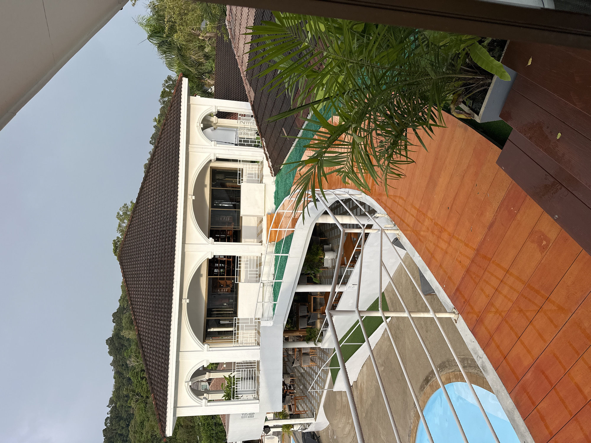 The image depicts a multi-level resort or restaurant building with a white exterior and dark tiled roofs, situated in Kathu, Thailand. A curved wooden walkway with an orange hue, possibly made of planks, leads from the foreground towards the building. This walkway is wet, with reflections visible on its surface, suggesting recent rain.
To the left of the walkway is a bright blue swimming pool with a rounded edge. The walkway is bordered by white railings, which also run along the edges of the building's various levels and ramps. Green artificial turf is installed on some of the ramps and walkways.
The building itself features arched openings on the upper level, revealing windows and balconies. Below this, a level with outdoor seating areas, including tables and chairs, is visible. Some potted plants are strategically placed around the outdoor seating and along the ramps.
In the background, lush green hills covered in trees rise, typical of the tropical landscape of Phuket. The sky is overcast, with a soft, diffused light, suggesting it is either early morning or late afternoon. A large potted palm plant is prominent in the foreground, partially obscuring the view of the walkway and the building.