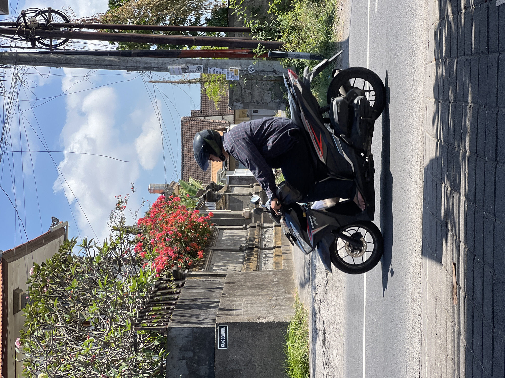 A man wearing a dark, long-sleeved, plaid shirt, dark pants, and a black helmet rides a black Honda scooter on a paved road. He is looking to his left and appears to be moving from left to right. The scooter has red and white accents. The road has a white line dividing the lanes. In the background, there is a grey concrete wall with a black sign that reads "GANG DEWI UMA". Behind the wall, there is a building with a red-tiled roof and vibrant pink bougainvillea flowers cascading over the wall. To the right of the road, there are utility poles with numerous wires tangled around them, and dense green foliage. The sky is blue with scattered white clouds. The scene appears to be in a residential area in Denpasar, Indonesia, characterized by the architecture and vegetation.