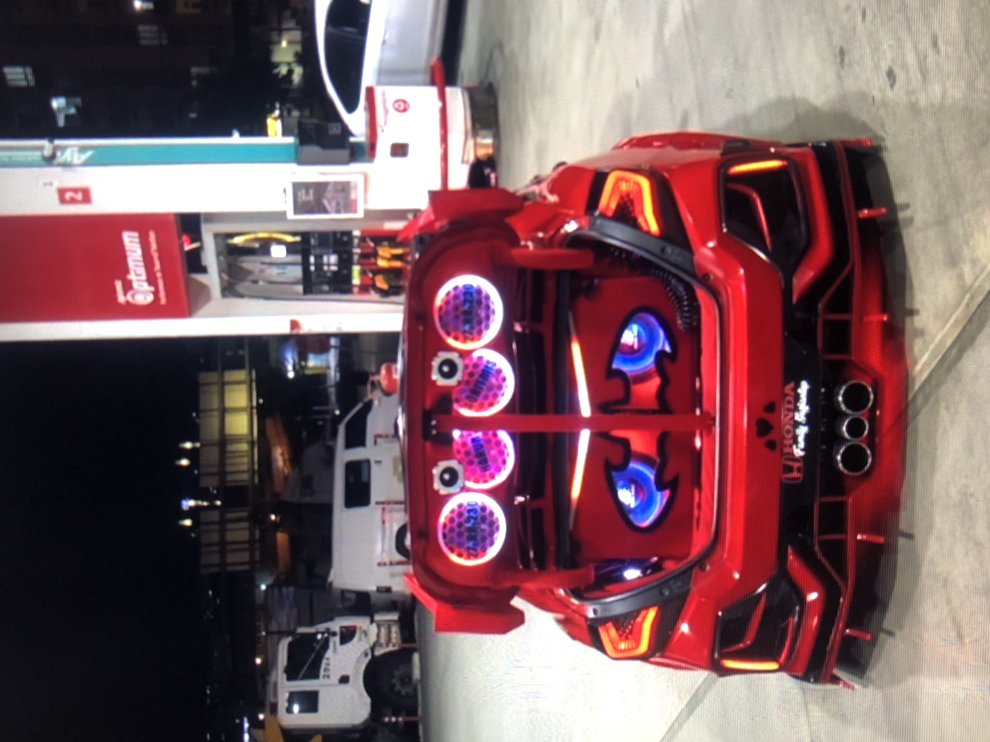 The image shows a red Honda vehicle with its trunk open, displaying a custom sound system and lighting. The car is parked at a gas station, specifically an Optimum station, indicated by the visible signage. Several other white vehicles are in the background, along with city lights suggesting an urban environment. Based on the stickers visible, including "Z-RM 230," and the Honda "Family Infinity" emblem, the location is determined to be Lhokseumawe, Indonesia. There are no visible people in the immediate foreground. The focus of the image is the car's modified trunk and its placement within a gas station environment.