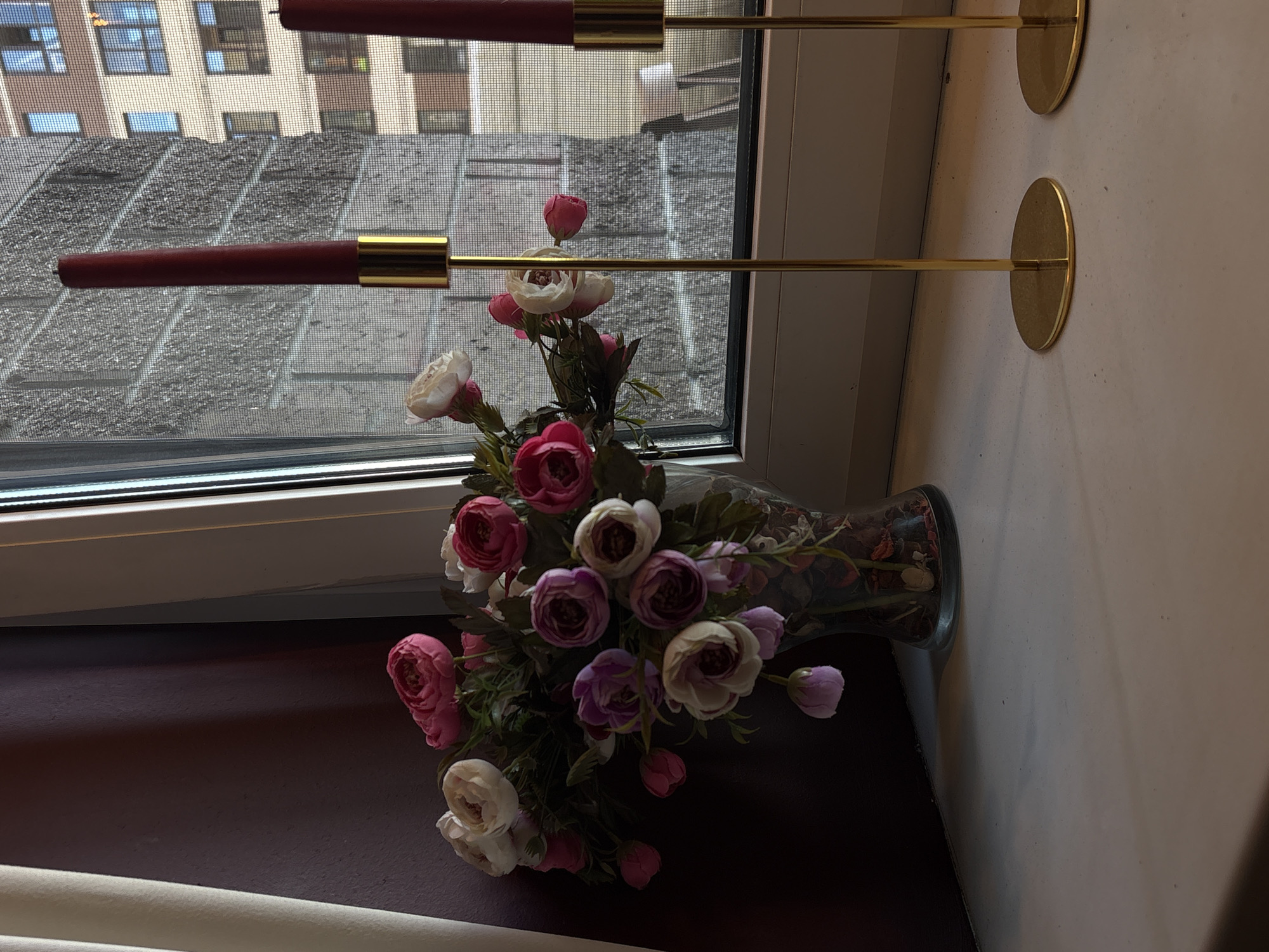 A vase of artificial flowers, predominantly pink and white, sits on a windowsill. Two red tapered candles in gold holders are positioned to the right of the vase. Through the window, a textured grey building with multiple windows is visible, suggesting an urban environment. The scene is likely indoors, with a curtain on the left side of the frame. There is no indication of people or any specific location within Saint Petersburg, Russia.