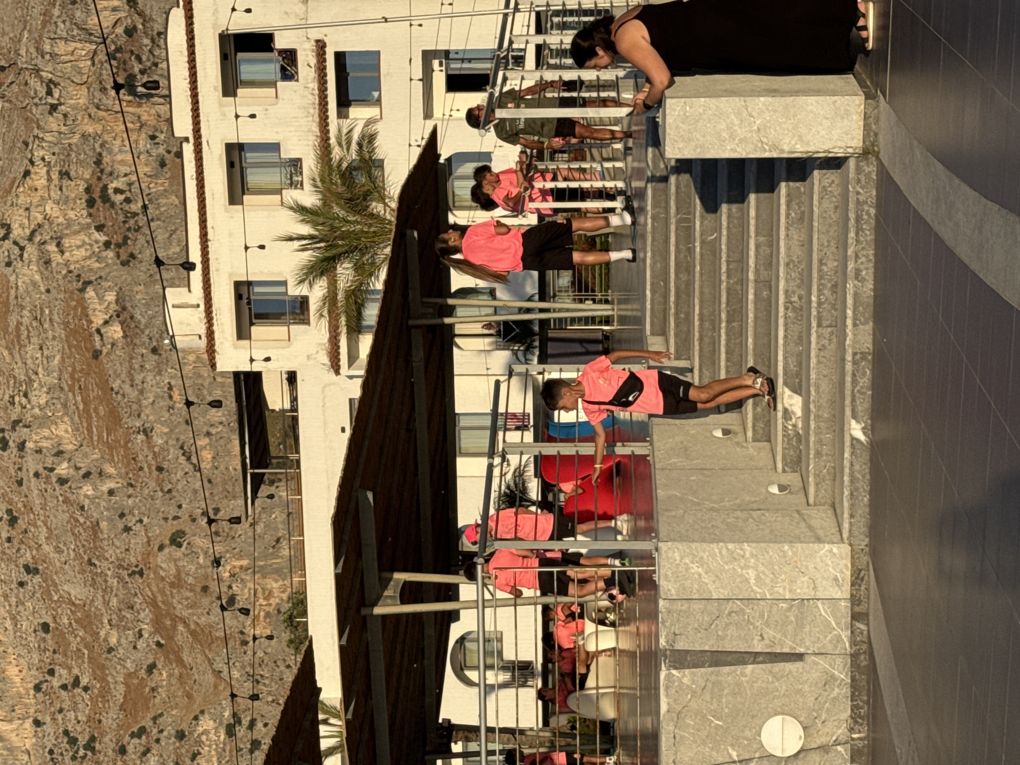 The image depicts a group of people, primarily young males wearing pink shirts and black shorts, on a multi-level outdoor staircase adjacent to a white building with balconies. Some individuals are standing, while others are sitting or lying down. One person is partially visible at the top, wearing a black dress and sandals. The building appears to be a hotel or residential complex, with several windows and a palm tree visible. The scene is set against a rocky hillside on the left. The lighting suggests it is late afternoon or early evening, with long shadows cast by the stairs and people. The overall atmosphere seems relaxed, possibly a break or a gathering. The location appears to be Isola delle Femmine, Italy, given the context. No visible text is present in the image.