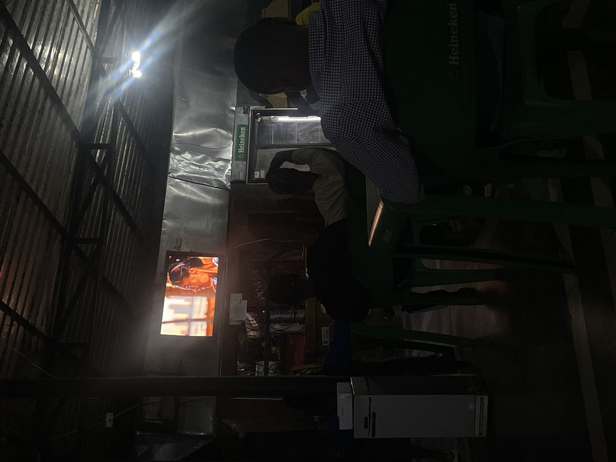 A dimly lit outdoor bar in Jos, Nigeria. Two people are sitting on green plastic chairs with Heineken logos. One person is wearing a checkered shirt. Another person is sitting at the bar near a Heineken branded refrigerator. A television on the wall displays a brightly lit image. The bar has a corrugated metal roof. There is a bright light under the roof. There are various objects and supplies near the bar area, including cardboard boxes and equipment under a table. The atmosphere appears casual.
