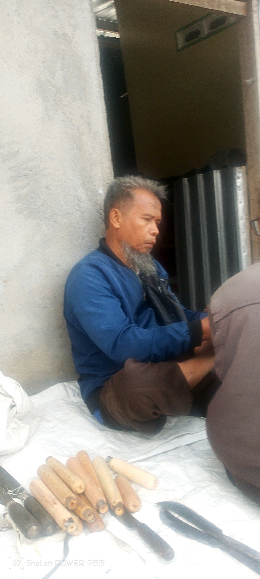 A side profile, medium shot captures a man with graying hair and a beard, wearing a blue jacket and brown pants, sitting on a white tarp outdoors. He is leaning against a textured concrete wall. In front of him on the tarp are several wooden-handled tools, possibly chisels or carving tools, as well as what appears to be a pair of tongs. The background consists of a corrugated metal structure and a wall with a small window. The lighting is overcast.