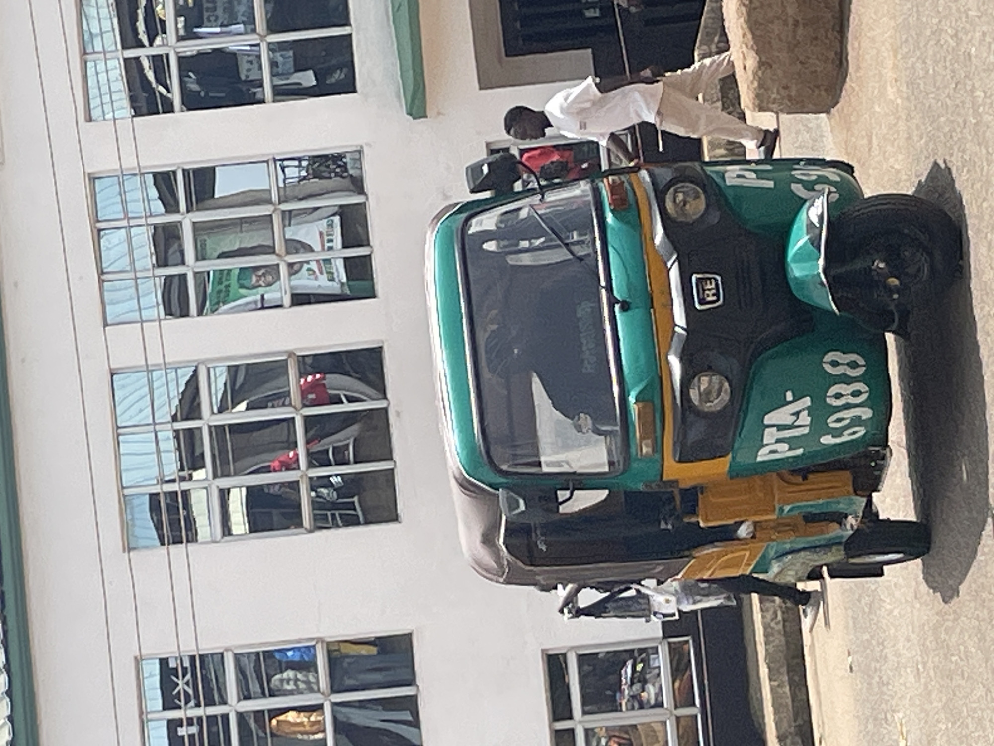In Bukuru, Nigeria, a three-wheeled auto-rickshaw, painted green and yellow, is parked on a paved road in front of a white building. The auto-rickshaw has "PTA-6988" written on its side. A man in a white t-shirt and tan pants is walking away from the auto-rickshaw to the right. In the background, the building has multiple windows with reflections of the sky and possibly interior objects. Some of the windows show what appear to be clothing or banners hanging inside. To the right of the building, a dark doorway is visible. A large, rough stone is situated on the right side of the frame.