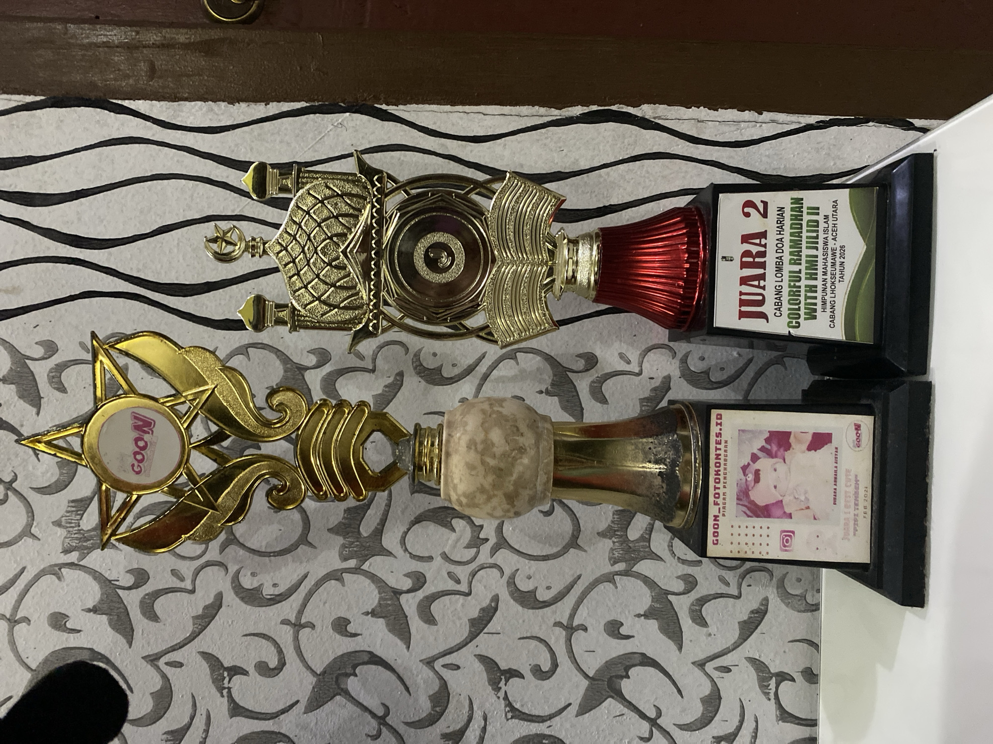 The image shows two trophies lying on a patterned surface. The top trophy has a gold-colored mosque design and a red-colored base. The base of the trophy indicates it's a "Juara 2" (2nd place) award for a "Colorful Ramadhan with HMI Jilid II" competition. The competition was organized by the Islamic Student Association in Lhokseumawe, Aceh Utara, in 2026. The second trophy has a gold-colored star design with the word "GOON" on it. The base of this trophy indicates it is a "Juara I Best Cute" (1st Place, Best Cute) for a competition called "GOON_FOTOKONTES.ID." The award was presented to Dinara Anwaila Aistan in February 2024.