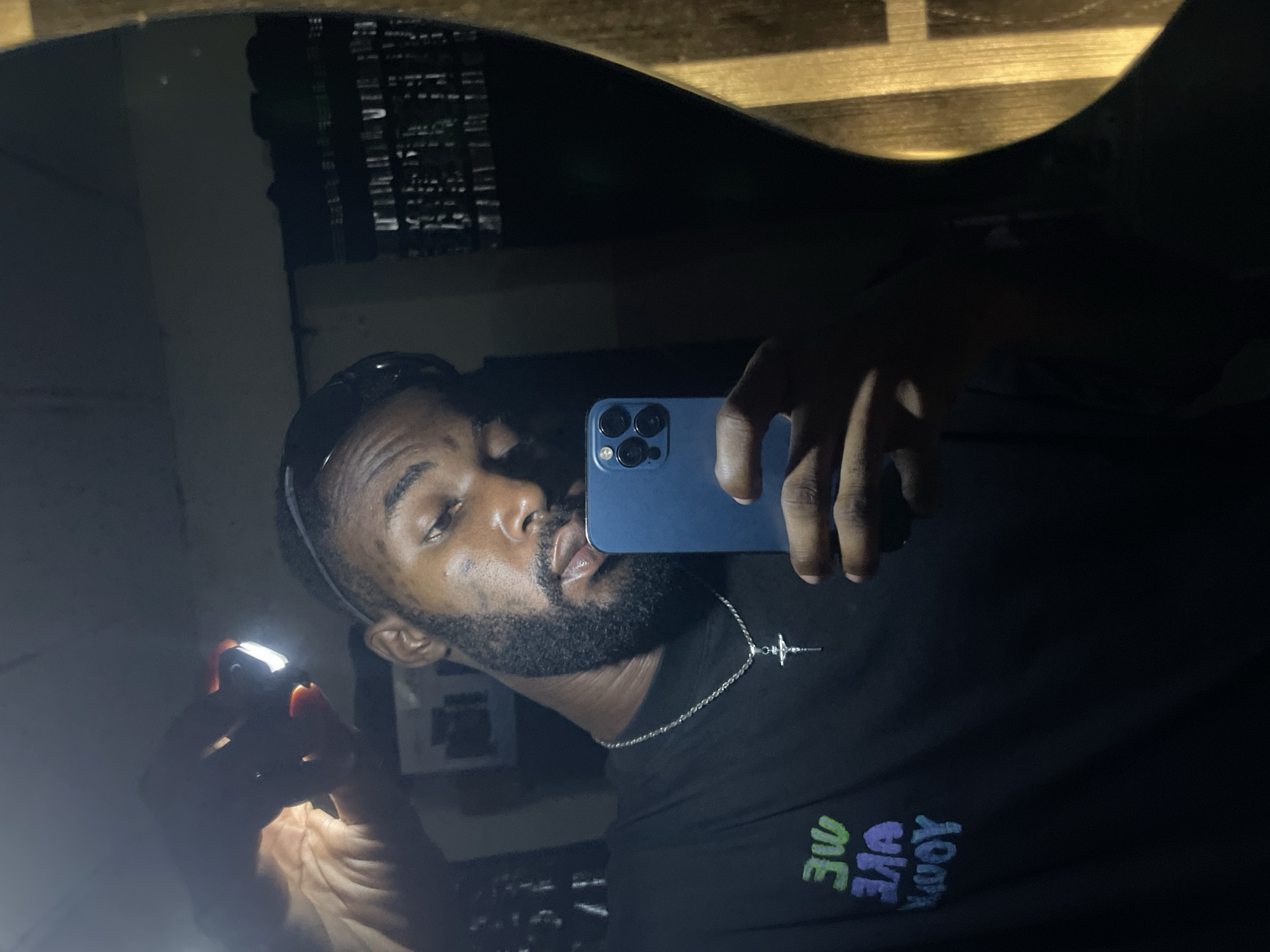 The image, taken in Lokoja, Nigeria, shows a man in a dimly lit room. He is holding a smartphone and a small flashlight, which illuminates his face and hand. He is wearing a black t-shirt with colorful text that appears to read "WE ARE YOUR". A silver chain with a crucifix pendant is visible around his neck. The background is dark and appears to be a wall with some reflective surfaces. The man is looking at his phone, suggesting he is taking a selfie or recording a video.