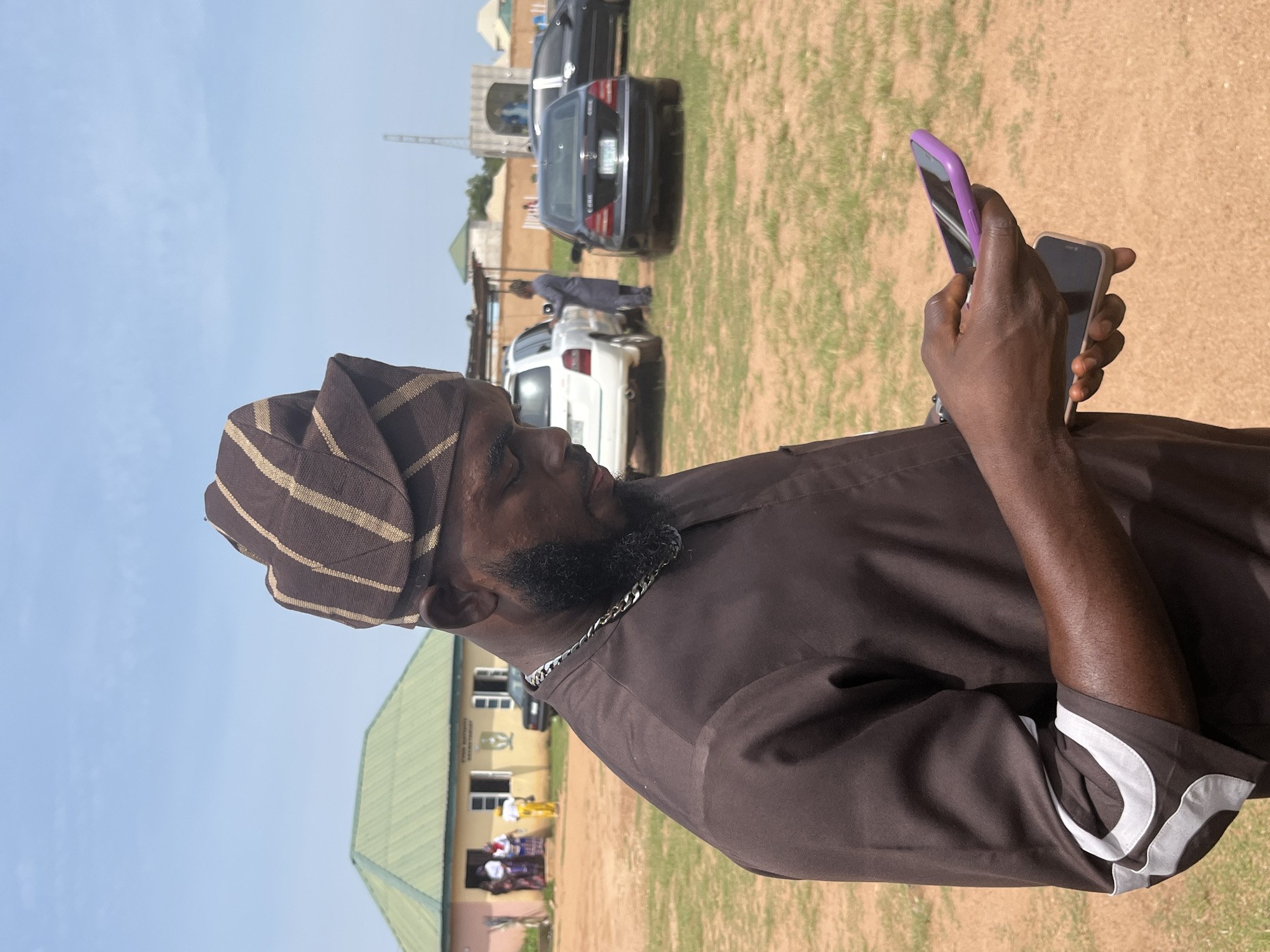 A man wearing a brown and gold striped cap and a brown tunic stands in an outdoor area with dry, sandy ground and sparse grass, looking down at two mobile phones in his hands. He has a dark beard and is wearing a silver chain around his neck. In the background, a yellow building with a green roof is visible, with a few people standing near the entrance. Several cars are parked in the background, including a white SUV, a black Mercedes-Benz sedan, and another black car. The sky is clear and blue, with a few wispy clouds. The location appears to be a public space, possibly a community center or a gathering place, in Jos, Nigeria.