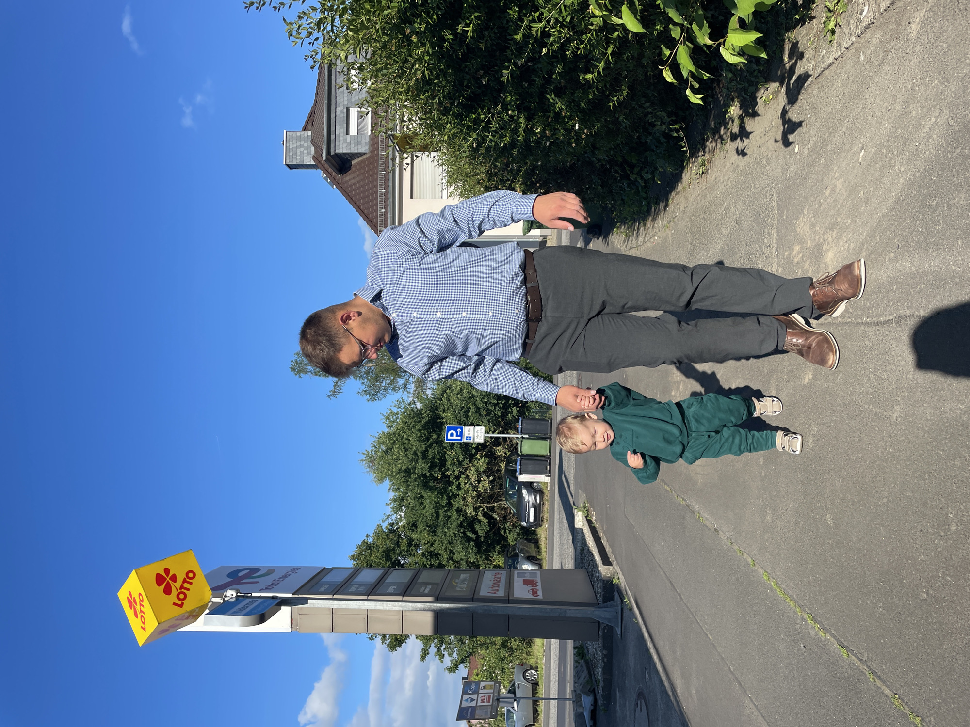 The image depicts a man and a toddler walking on a sidewalk in front of a Total gas station in Kohden, Germany. The man, wearing a blue checkered shirt, dark pants, and brown shoes, is holding the toddler's hand. The toddler, dressed in a green tracksuit and sandals, is looking down and appears to be walking independently. The background features a gas station sign with a yellow Lotto cube, trees, and a residential building. The sky is clear and blue, with a few white clouds visible. The lighting suggests it is a sunny day.