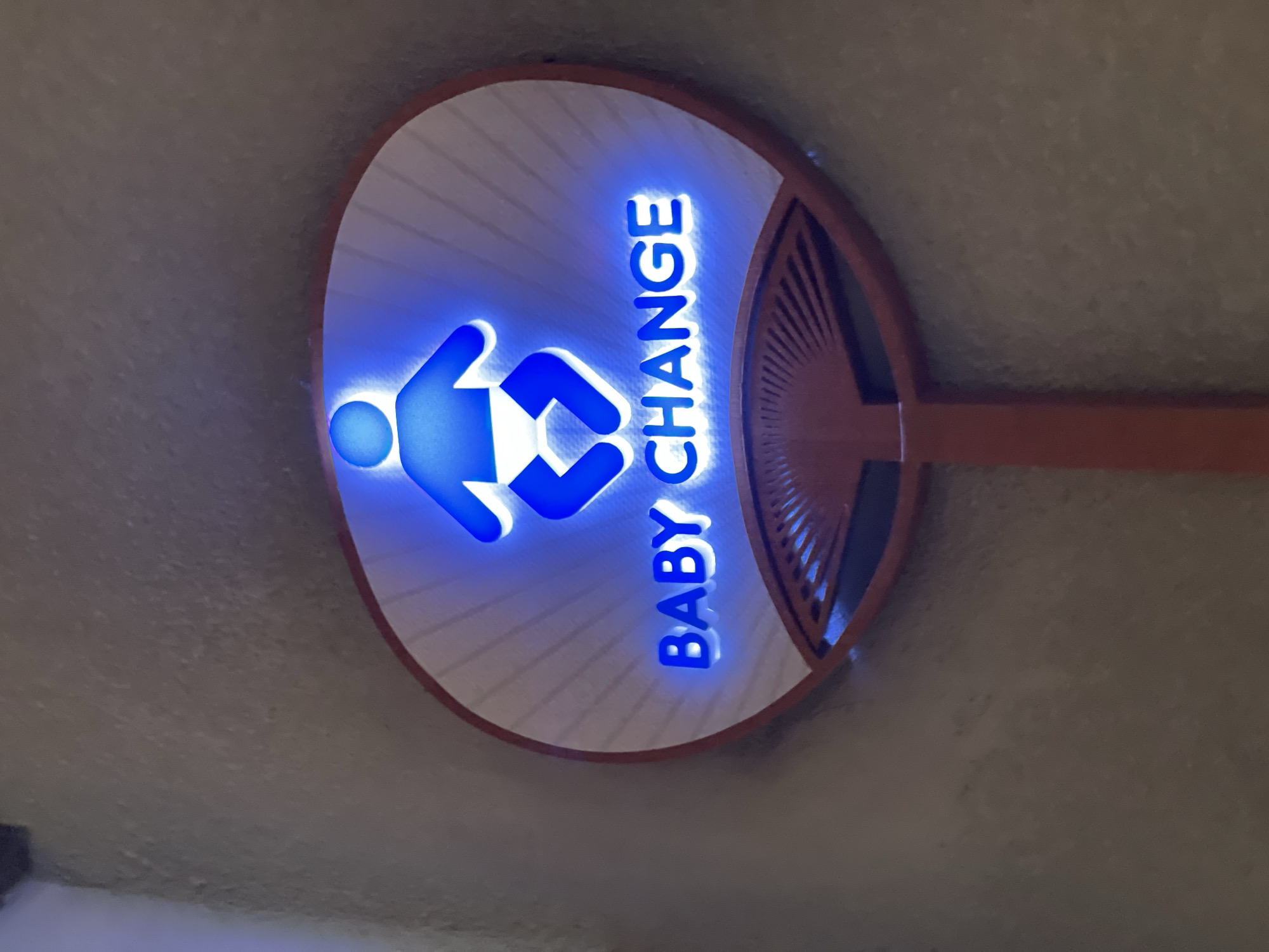 The image shows a circular, fan-shaped sign with a blue illuminated baby icon at the top and the words "BABY CHANGE" in blue illuminated letters below it. The sign is mounted on a red pole and is set against a textured, beige wall. The sign appears to be an indicator for a baby changing facility. The context of Pattaya City, Thailand, is not visually evident in the provided image, but the sign's function suggests it is located in a public place such as a mall, restaurant, airport, or public restroom within Pattaya City.