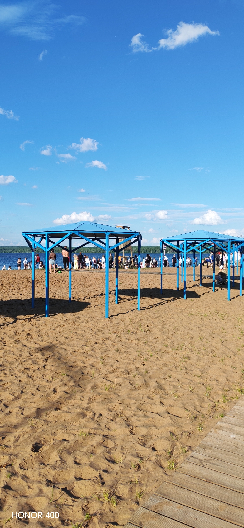 The image depicts a sunny day at a sandy beach, likely in Chaikovsky, Russia, near the Kama River. Two blue, hexagonal gazebos are prominent in the foreground, casting shadows on the sand. In the midground, a body of water, presumed to be the Kama River, stretches across the horizon, with a tree-lined shore in the distance. A crowd of people are gathered along the water's edge and on the beach, some appearing to be sunbathing or socializing. A wooden walkway is visible on the right side of the frame, leading away from the beach. The sky is a vibrant blue, dotted with fluffy white clouds. A lifeguard tower is situated behind the first gazebo. The overall scene suggests a popular recreational area on a warm summer day.