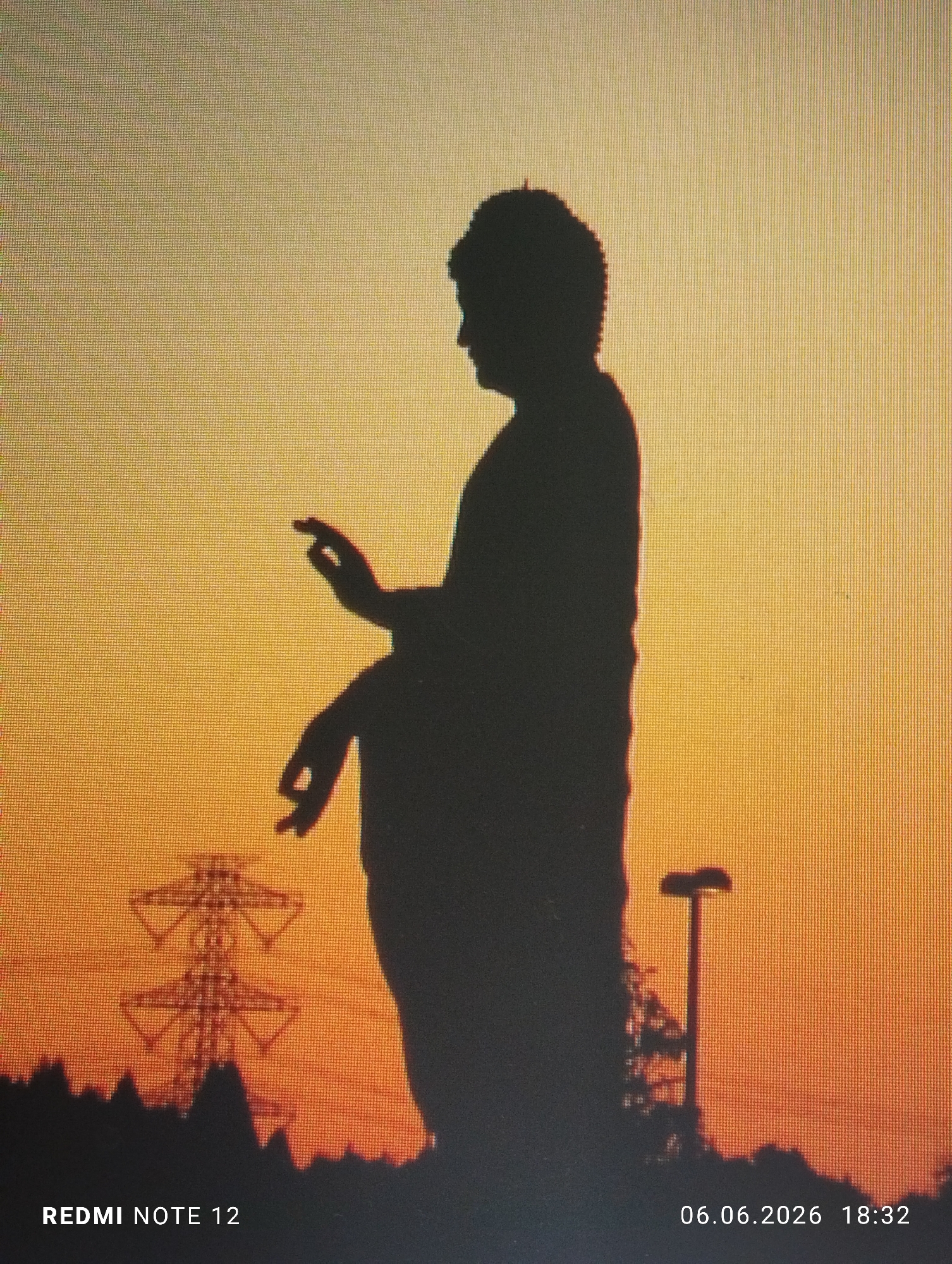 The image is a digital photograph of a screen displaying a large, dark silhouette of a standing statue, resembling a religious or meditative figure, against a vibrant sunset sky. The figure's right hand is raised with the thumb and index finger touching, forming a circle, while the left hand is lowered and slightly bent. The head features a textured or coiled outline. The sky exhibits a gradient from golden yellow at the top to deep orange and reddish tones towards the horizon. In the background on the left, a prominent electrical transmission tower is silhouetted. On the right, a tall, pole-mounted street light with a distinctive top is visible as a silhouette. The foreground consists of an undulating dark silhouette of terrain or foliage. The entire image displays a visible pixel grid or moire pattern, characteristic of a photograph taken of a digital display. Embedded text at the bottom left reads "REDMI NOTE 12," indicating the original capture device, and at the bottom right, "06.06.2026 18:32," indicating the date and time of the original image capture. The location is Unknown, Unknown.