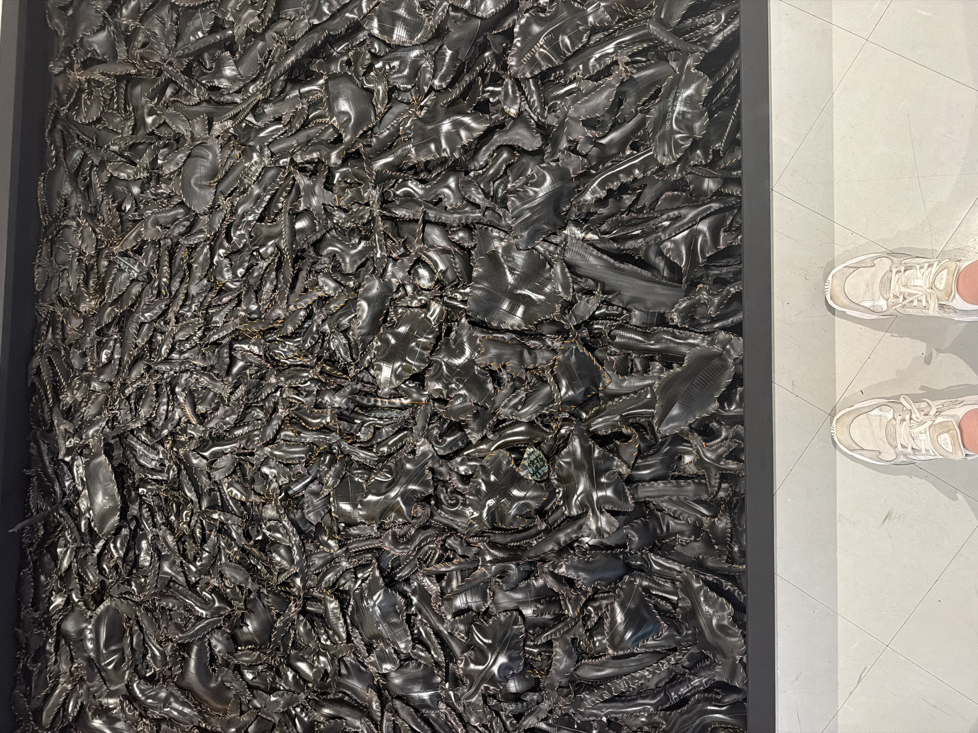 An overhead, full-frame shot shows a massive art installation made of black, shiny, textured material, possibly recycled rubber, pieced together with visible stitching. The material is arranged in a dense, organic, and somewhat chaotic pattern, filling the entire frame from edge to edge, with some pieces appearing to overlap and fold over one another. The surface has a metallic sheen, reflecting ambient light and creating highlights and shadows that add depth and texture. In the foreground, at the bottom of the image, a person's feet are visible, wearing a pair of light-colored athletic shoes. They are standing on a light gray tiled floor, with faint lines marking the tile grout. The art installation is framed by a dark, horizontal border at its base. The scene appears to be an indoor exhibition space, possibly in a gallery or museum. The city of Bangkok, Thailand, is not directly depicted in the image, nor is there any information in the visual data to confirm its location.