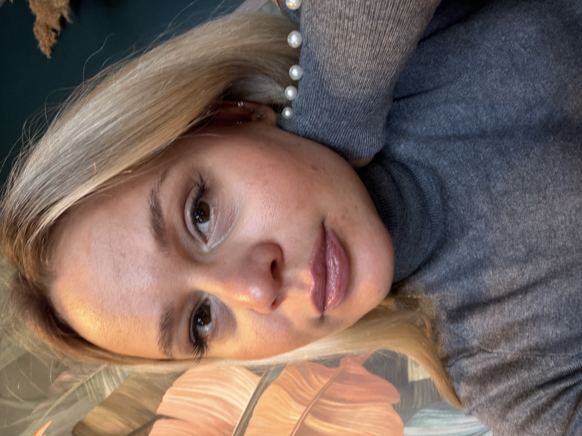 A blonde-haired woman is shown in a close-up portrait. She has brown eyes and fair skin. She is wearing a grey turtleneck sweater and a string of pearls around her neck. The background is out of focus, but there is a plant with peach-colored leaves visible. The city of Sarapul, Russia is not discernable from the visual data.