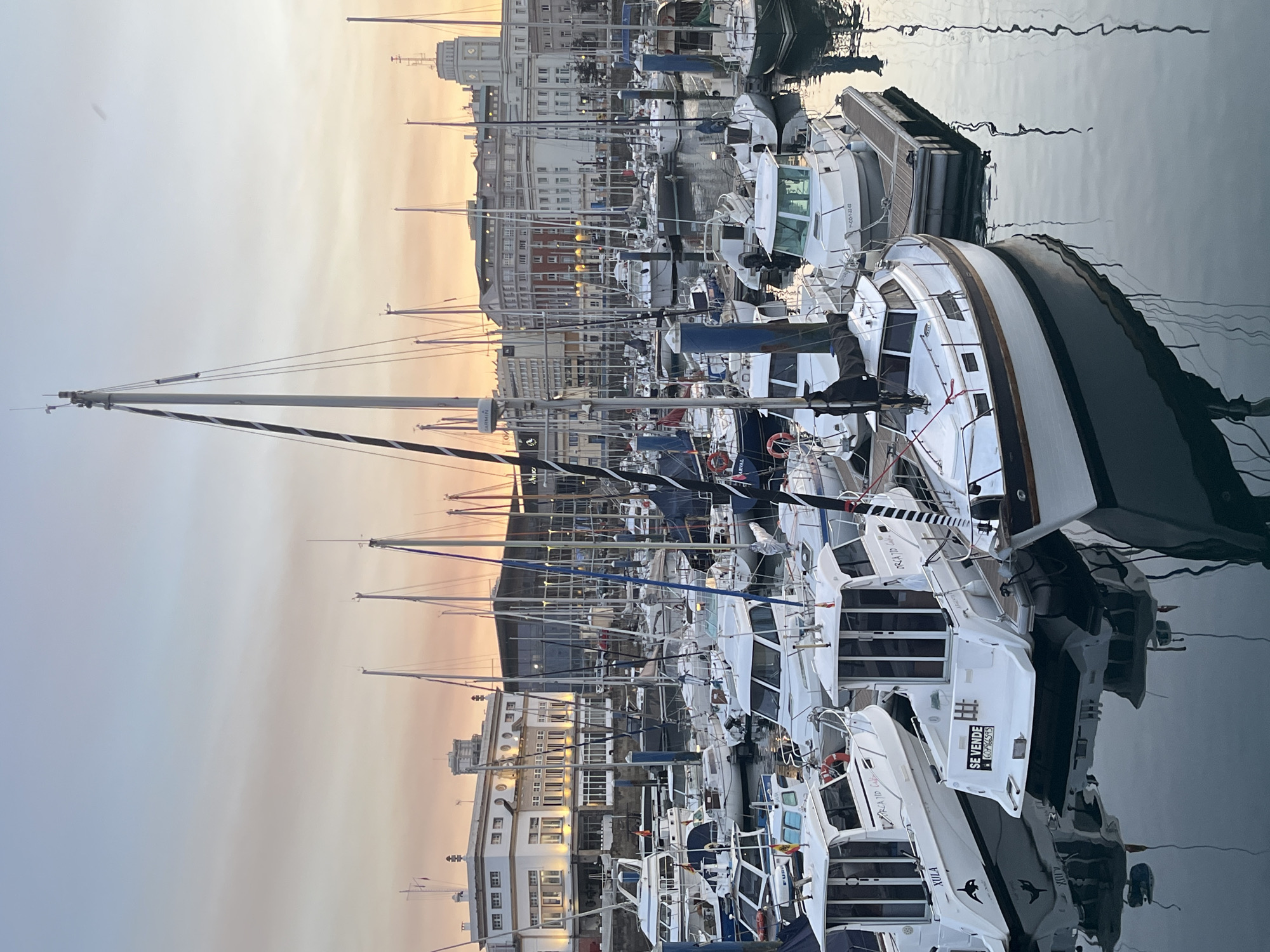 This is a photograph of a marina in A Coruña, Spain, taken at dusk. The sky is a soft gradient of blue and orange, indicative of sunset. Numerous sailboats and motorboats are docked, their masts reaching towards the sky. Some of the boats are white, while others have darker hulls. Reflections of the boats and their masts are visible on the calm water of the harbor. In the background, several multi-story buildings are visible, some with lit windows, suggesting urban development surrounding the marina. One building appears to be a distinct white structure with arched windows. The overall atmosphere is tranquil and serene, characteristic of a marina at the end of the day.