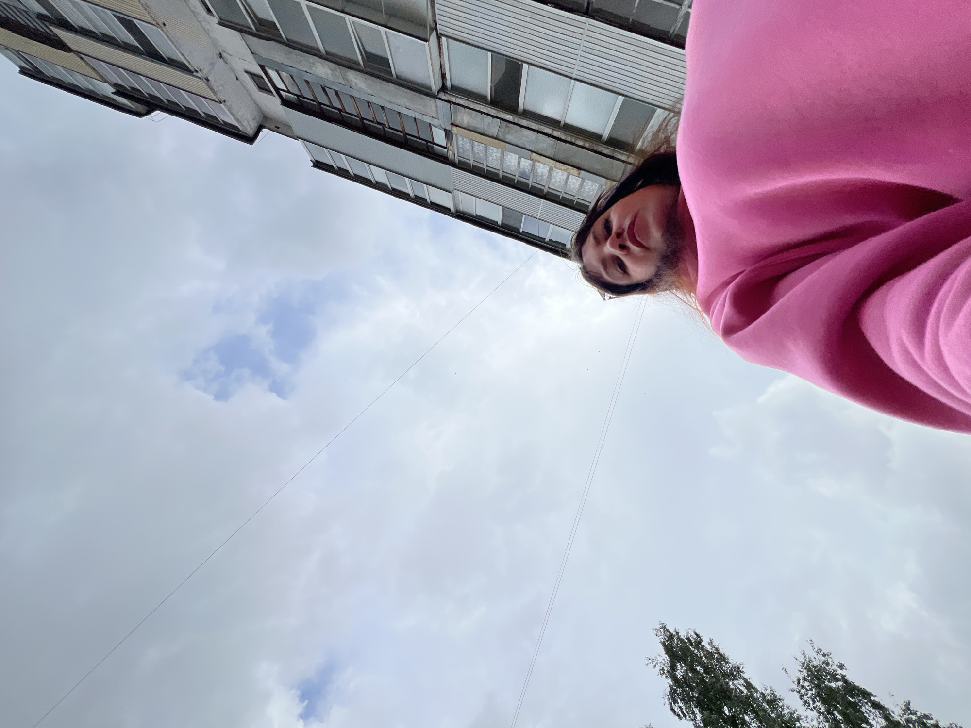 An outdoor, low-angle shot captures a person looking down at the camera, framed against a cloudy sky and an apartment building. The subject, likely female, is partially visible at the upper right, wearing a vibrant pink hoodie. Her face is directed downwards with a neutral to slightly smiling expression, her dark hair falling around her shoulders.
In the background, a multi-story building with numerous windows and balconies occupies the upper left quadrant of the frame. Its architecture suggests a utilitarian style, common in residential areas in places like Noviy, Russia. The sky is predominantly overcast with soft grey clouds, indicating a cloudy or partly cloudy daytime setting. Thin, dark wires crisscross the middle of the sky, stretching diagonally across the frame. At the bottom right, the tops of several trees with dense green foliage are visible, adding a natural element to the urban-dominated scene. No discernible text is visible in the image.