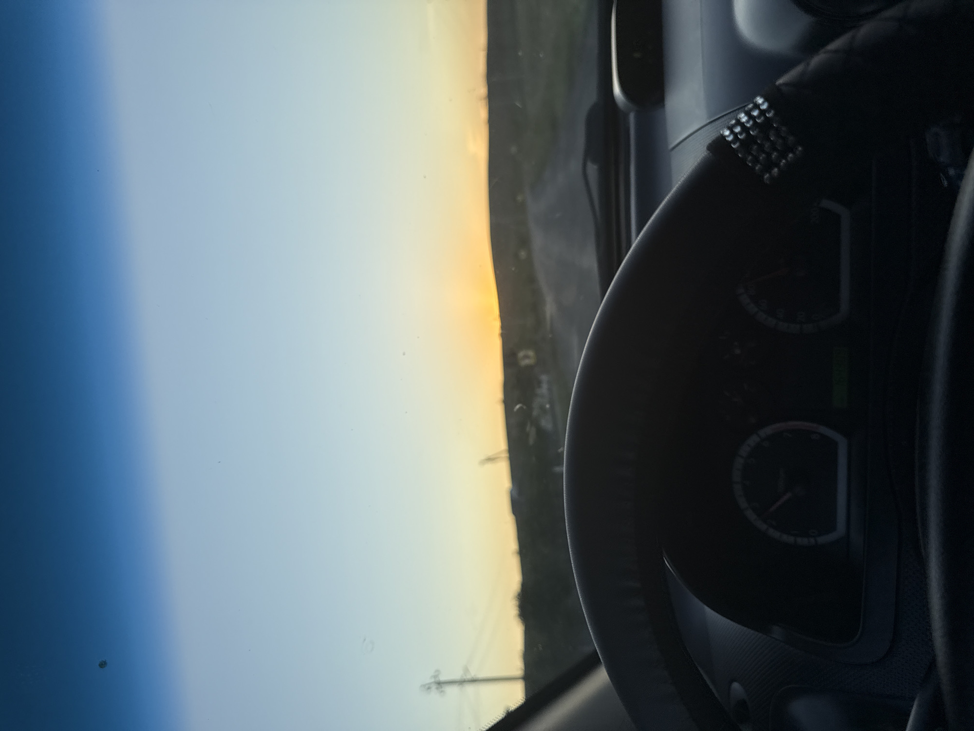 The image, taken from inside a vehicle, captures a view through the windshield of a rural landscape during sunset. A steering wheel is prominently visible in the foreground, adorned with a decorative band of rhinestones. The dashboard displays a partially obscured speedometer. The road ahead appears unpaved, winding through a gently rolling terrain. In the distance, the sun is setting, casting a warm glow on the horizon. A utility pole with electrical wires stands to the left of the frame. The sky transitions from a deep blue overhead to a pale yellow and orange near the horizon. The location is identified as Cioc-Maidan, Moldova.