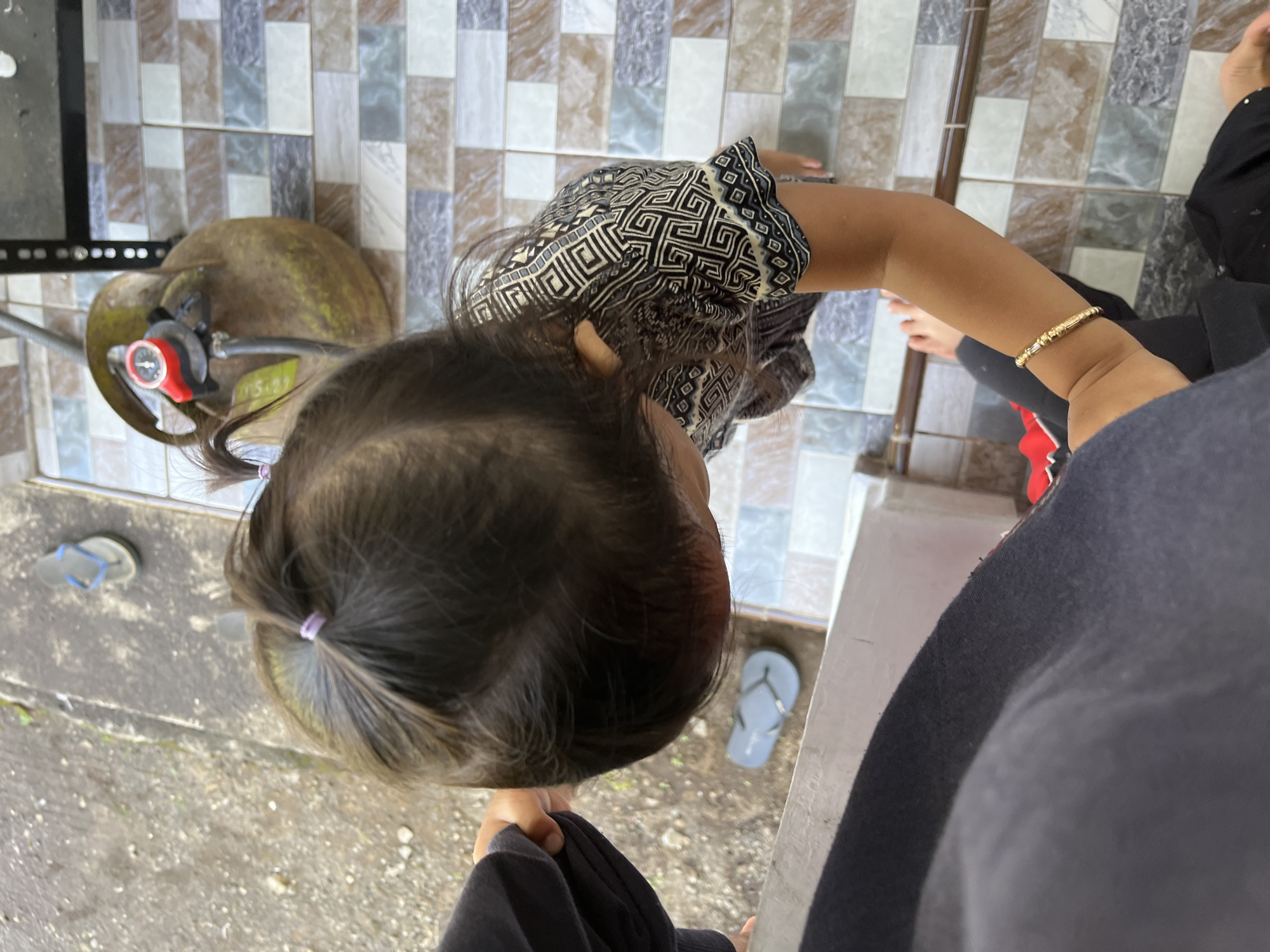 A young child with dark hair styled in two pigtails is captured from a high-angle perspective, looking down at the ground. The child is wearing a black and white patterned shirt and black pants, with a thin gold bracelet on their right wrist. They are standing on a tiled floor with a pattern of beige, brown, and grey squares. To the left of the child, a person wearing a dark grey garment is partially visible, their hand extended towards the child. In the background, a rusty-colored gas cylinder with a red regulator is connected to a pipe. The ground outside this area appears to be a rough, grey surface, with a single blue flip-flop visible near the edge. The scene is outdoors, with a glimpse of a railing or edge in the foreground.