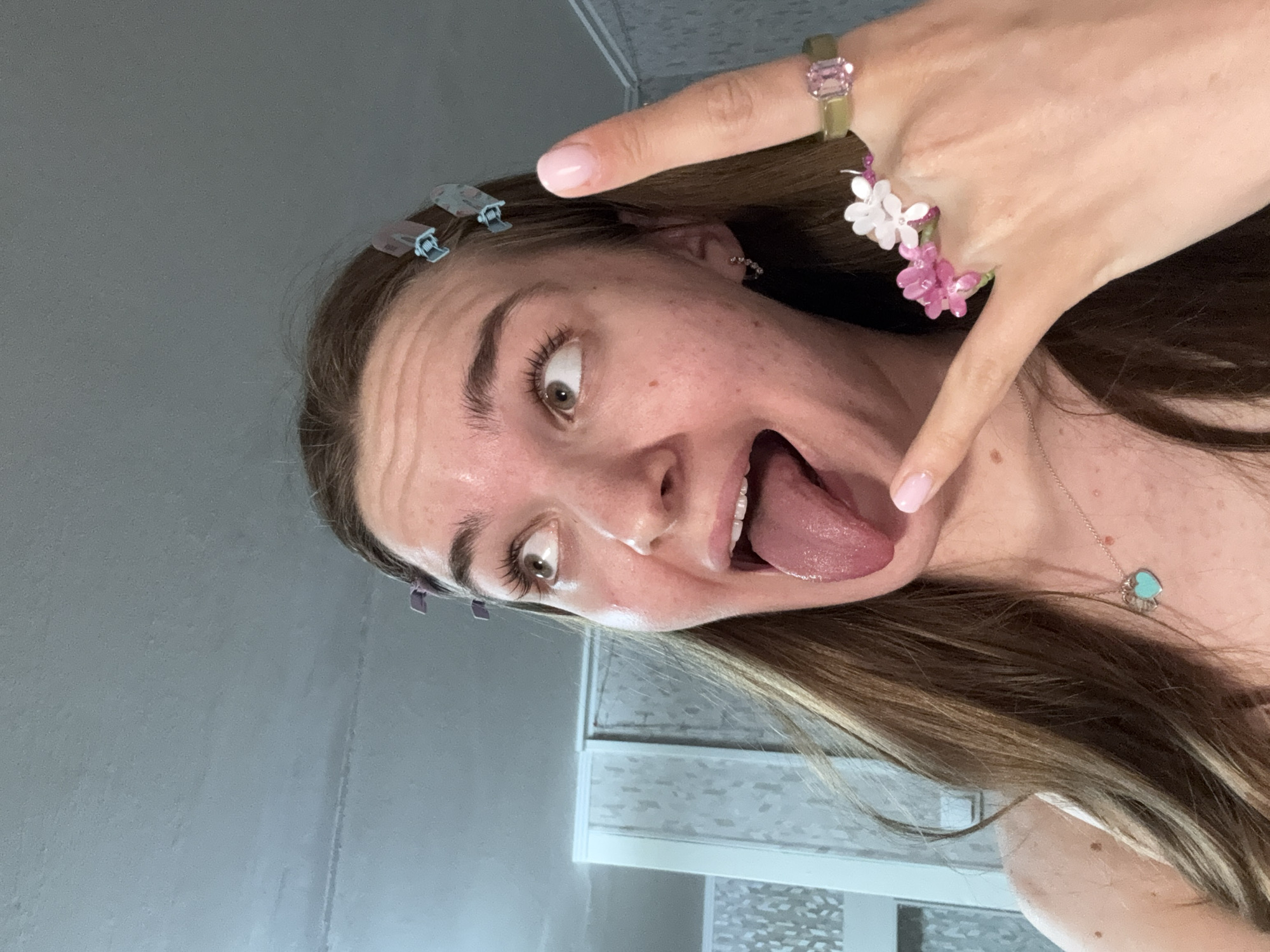 A woman with light brown hair and fair skin is posing for a selfie indoors. She is sticking her tongue out with wide eyes and a playful expression. She is wearing blue hair clips in her hair, a gold ring with a pink gemstone, and a ring with pink and white beaded flowers. She also has on a delicate necklace with a heart-shaped pendant. The background appears to be a bathroom with a gray wall and white tiled surfaces. The location in Perm, Russia cannot be verified.