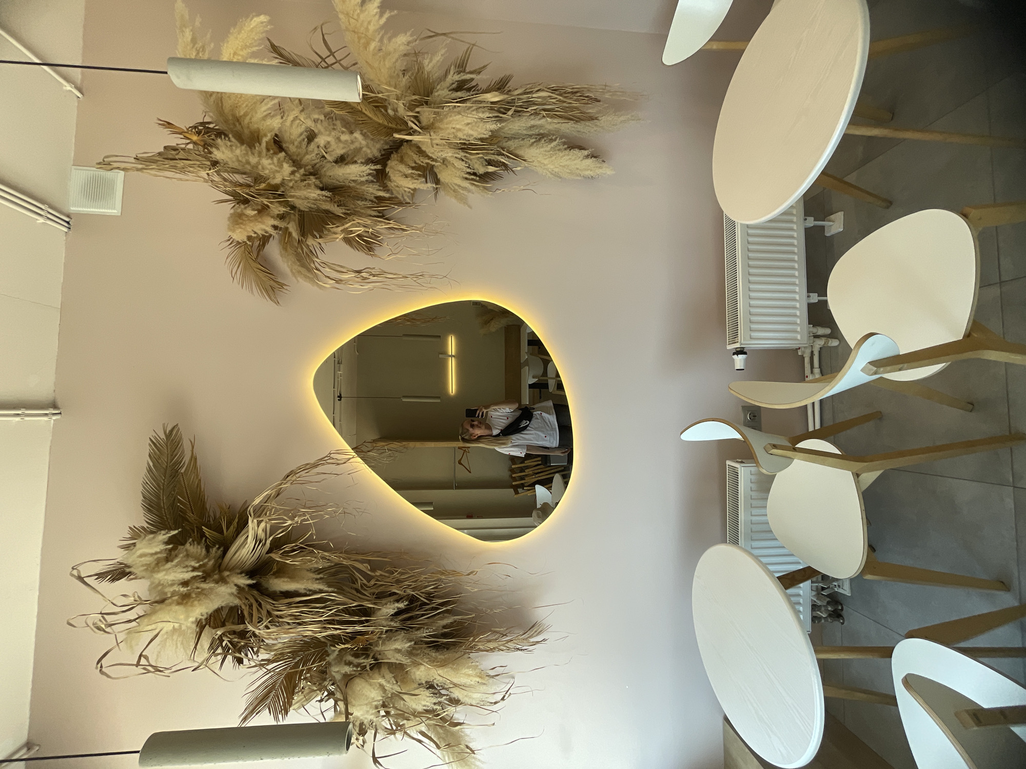 This image captures the interior of what appears to be a modern cafe or restaurant, located in Vladivostok, Russia. The walls are painted a soft, pale pink or beige color, contributing to a calm and inviting atmosphere.
The primary subjects include contemporary furniture, decorative dried plants, and a distinctive mirror. On the left side of the wall, two large arrangements of dried pampas grass and other natural-toned dried foliage are mounted, serving as prominent decorative elements. Each arrangement is illuminated by a minimalist, white cylindrical pendant light.
In the center of the image, a uniquely shaped mirror with a warm, glowing LED backlight stands out. The mirror reflects a person, likely the photographer, taking a mirror selfie with a smartphone. The person has light-colored hair, is wearing a white short-sleeved top, and a black fanny pack or sling bag across their chest. Other reflected elements inside the mirror include a wooden clothes hanger and parts of the room beyond the main space, possibly revealing more minimalist design and wooden architectural features.
To the right of the mirror, several light-colored, round-topped tables are arranged with matching chairs featuring curved seats and backs, all supported by light wooden frames. Below one of these tables, a white radiator with visible pipes is installed against the wall.
High on the upper left wall, a white square ventilation grate is visible, along with some white conduits or cables running neatly along the ceiling line.
The overall aesthetic blends modern minimalism with natural, earthy decor. The main activity depicted is the act of taking a mirror selfie. There is no visible text in the image. The time of day is not discernible, but the interior lighting suggests a well-lit indoor environment.