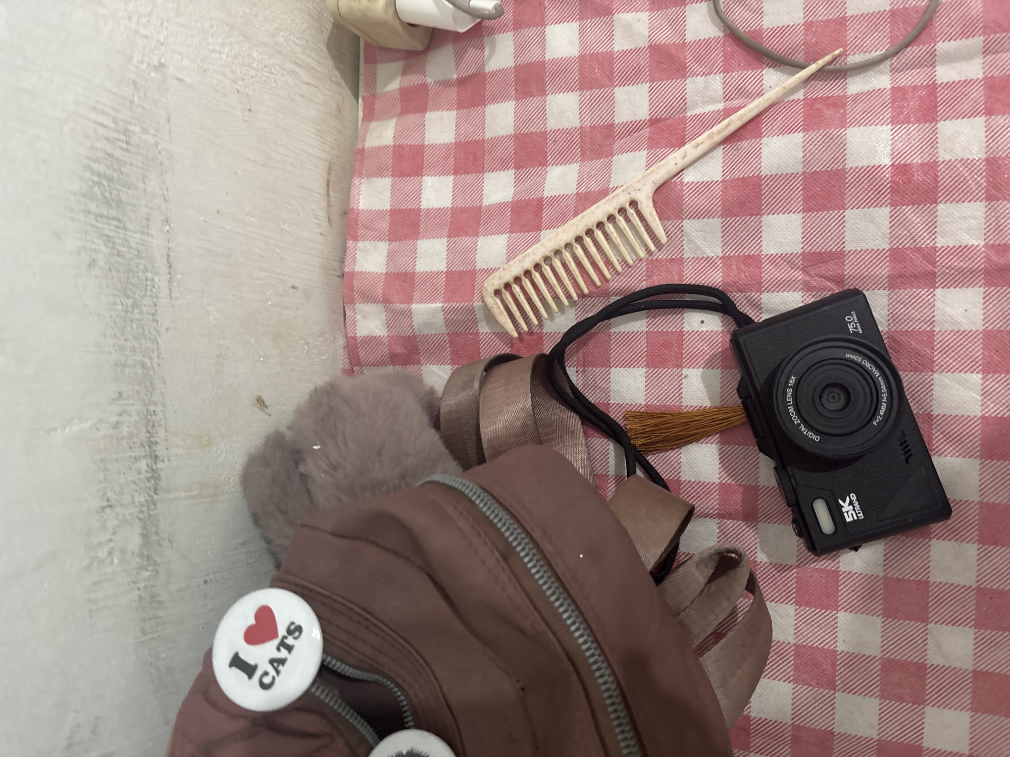 A close-up, top-down shot shows a collection of items arranged on a pink and white checkered fabric. The items include a brown backpack with a zipper and two circular pins attached to it, one of which reads "I ❤️ CATS." A fluffy, grey item is partially visible behind the backpack. A beige comb with wide teeth lies to the right of the backpack. In the foreground, a black digital camera is placed on the fabric. The camera has text labeling it as "5K ULTRAHD," "DIGITAL ZOOM LENS 18X," and "75.0 MEGA PIXELS." A thin, brown tassel is attached to the camera. The background appears to be a white, textured wall with faint gray markings. The overall scene suggests a personal collection of belongings, possibly in a domestic setting. There is no discernible location in the city of North Jakarta, Indonesia, as this is a still image without contextual clues related to geography or specific landmarks.