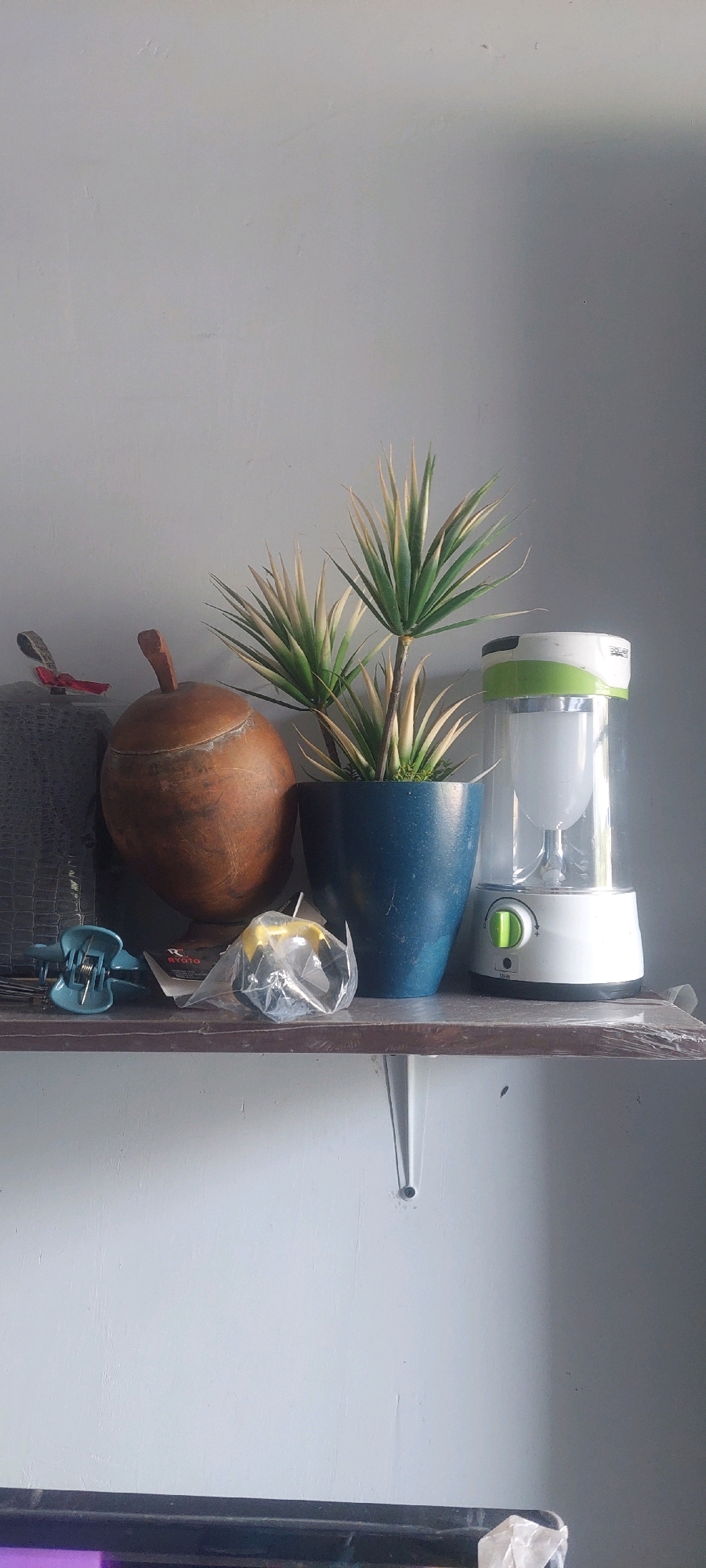 The image depicts a shelf with various objects arranged against a light gray wall. On the left, there is a dark gray textured object with a red ribbon tied around its top. Next to it is a round, brown, gourd-like object with a wooden handle. To the right of the gourd is a potted plant with spiky green and white leaves, housed in a blue ceramic pot. Further to the right is a white and green portable lantern with a transparent cylinder. On the shelf in front of these items are two blue hair clips and a small, clear plastic-wrapped object containing what appears to be a yellow and black item. The shelf itself is a dark brown wood, supported by white metal brackets mounted on the wall. The scene is indoors, likely in a home or storage area, with natural light casting shadows on the wall and objects. The image does not contain any contextual information that would allow for determining its location in Lumajang, Indonesia.