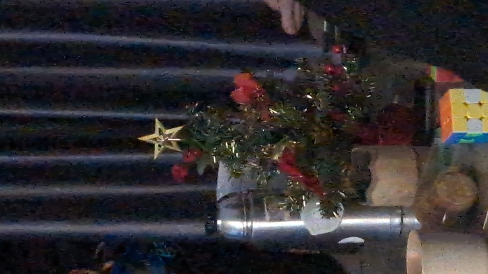 This image depicts an indoor environment in Thành phố Đà Lạt, Vietnam, characterized by low light and visible digital noise.
The central object is a small artificial Christmas tree, adorned with green foliage, red decorative elements (appearing as small baubles or clusters), and gold tinsel. A gold star serves as the tree topper. The tree rests within a visible red pot.
To the left of the Christmas tree, a tall, cylindrical stainless steel object, possibly a thermos or insulated container, stands on the surface. Next to this, closer to the foreground, is a light-colored planter, appearing to be ceramic or concrete, featuring a wavy or scalloped upper rim and containing green foliage distinct from the Christmas tree.
In the bottom left corner, a partial view of a light-colored, possibly ceramic, mug is visible. In the bottom right corner, a multi-colored Rubik's Cube is present, displaying blue, yellow, and orange faces, with some white squares visible.
All objects are arranged on a flat, dark, and somewhat reflective surface, likely a table. The background consists of dark, vertically pleated or striped material, consistent with curtains or blinds. No individuals are visible, and no actions or interactions are depicted within the static scene.