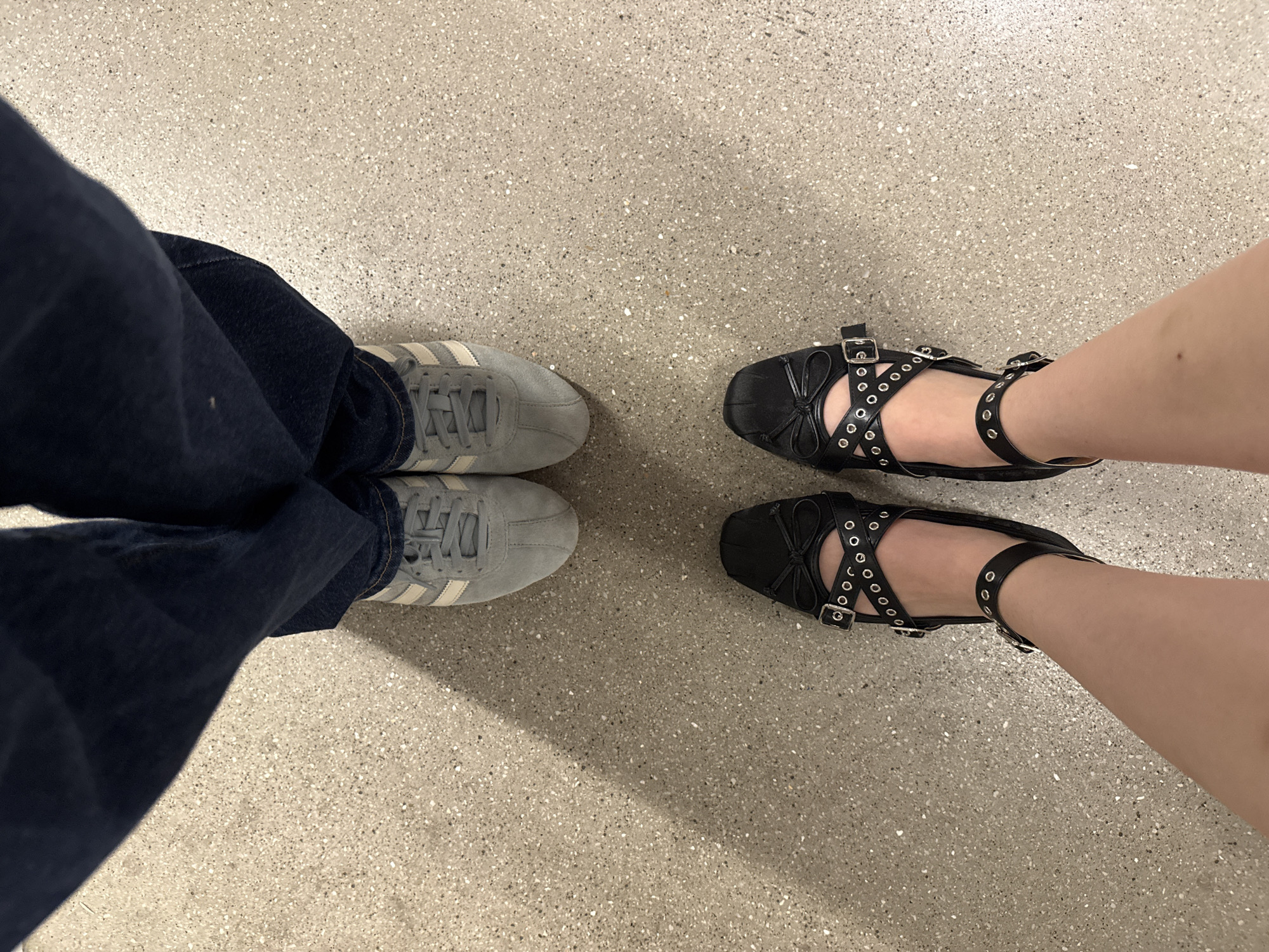 The image shows a view from above of two pairs of feet standing on a speckled light-grey surface. On the left, a pair of feet is wearing blue jeans and grey sneakers with white stripes. On the right, a pair of feet is wearing black ballet-style flats with multiple straps and eyelet details. The setting appears to be an indoor space with artificial lighting, possibly in Mexico City, Mexico.
